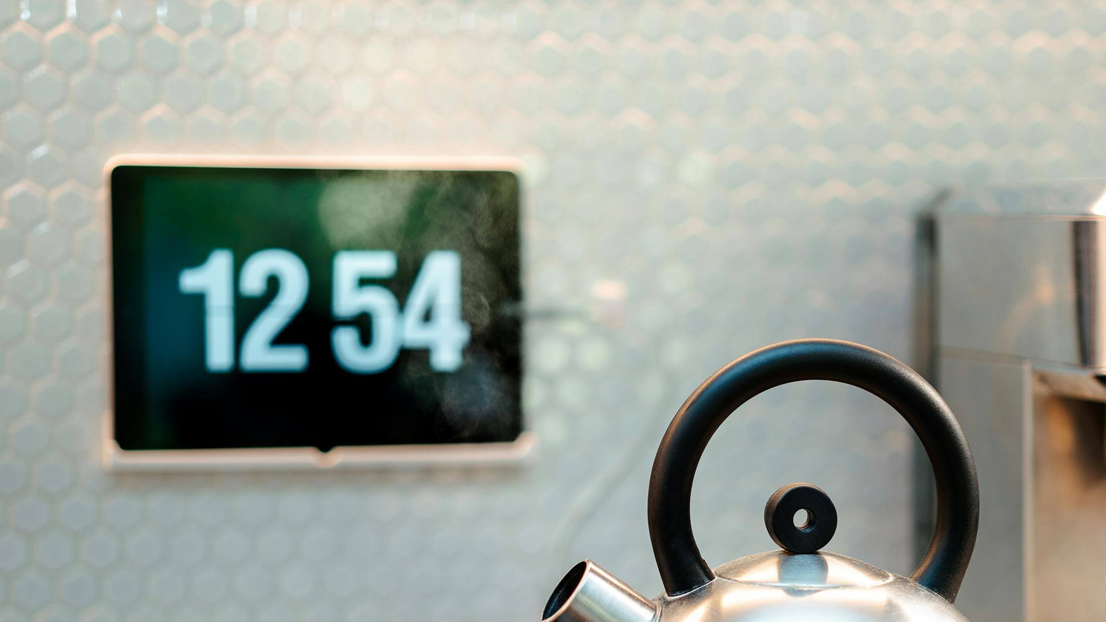 Digital clock showing 12:54 with steam rising from a stainless steel kettle spout in the foreground