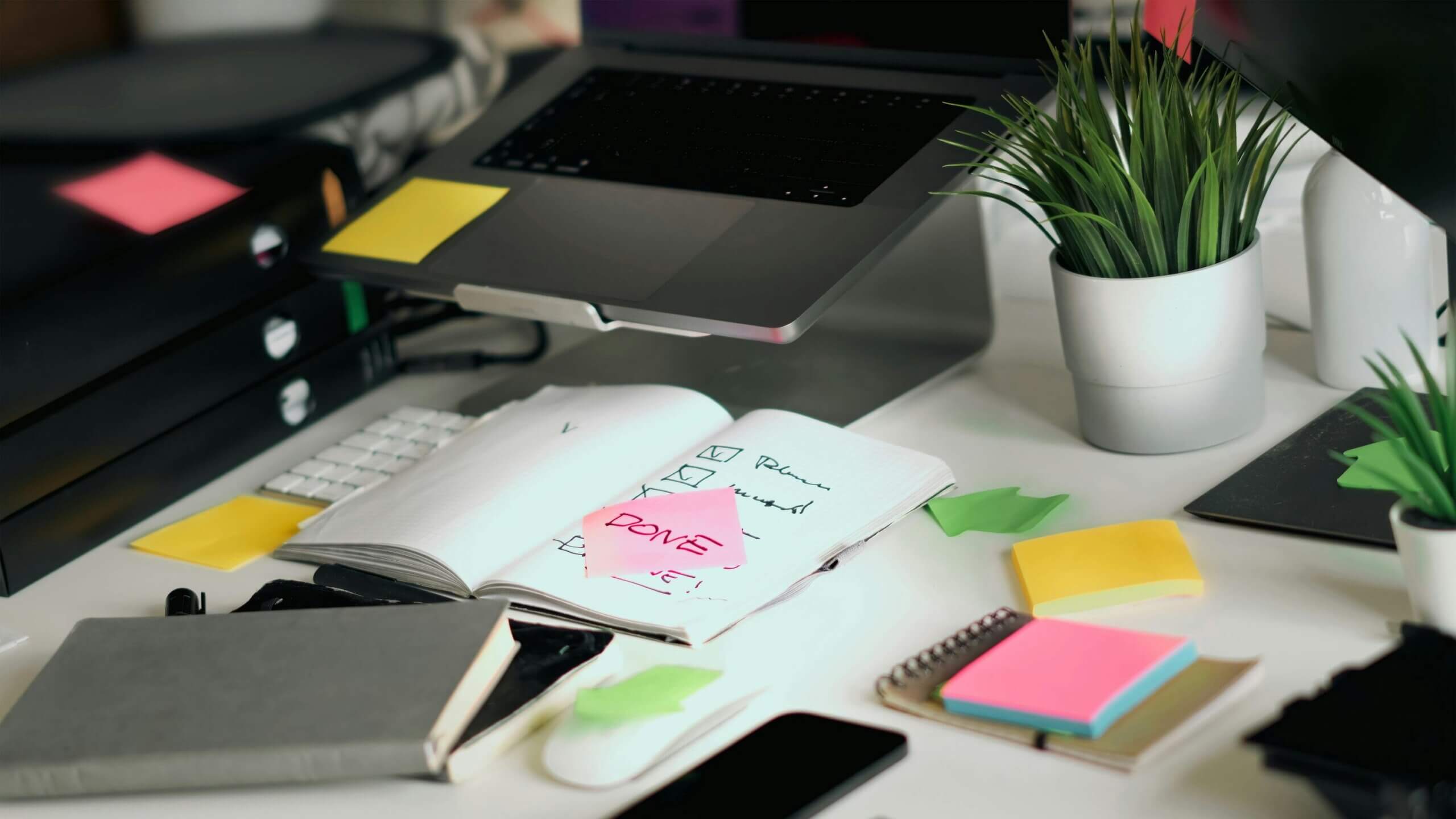 Desk with open notebook marked "DONE" on a pink sticky note, laptop, keyboard, phone, and potted plants.