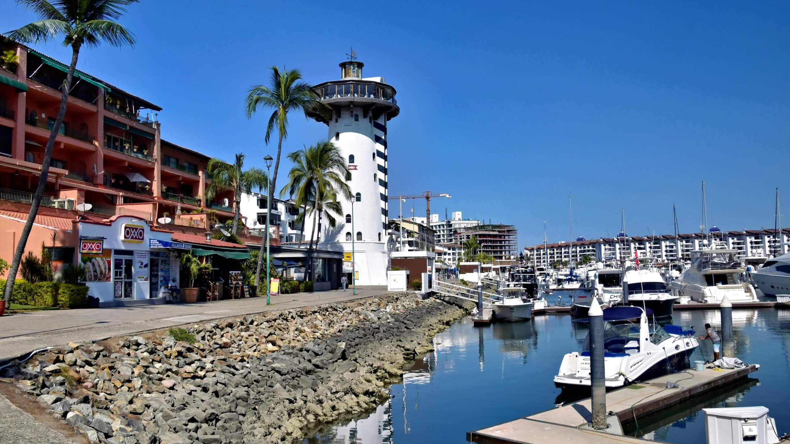 Marina with boats docked near a white lighthouse and an OXXO convenience store under clear blue sky.