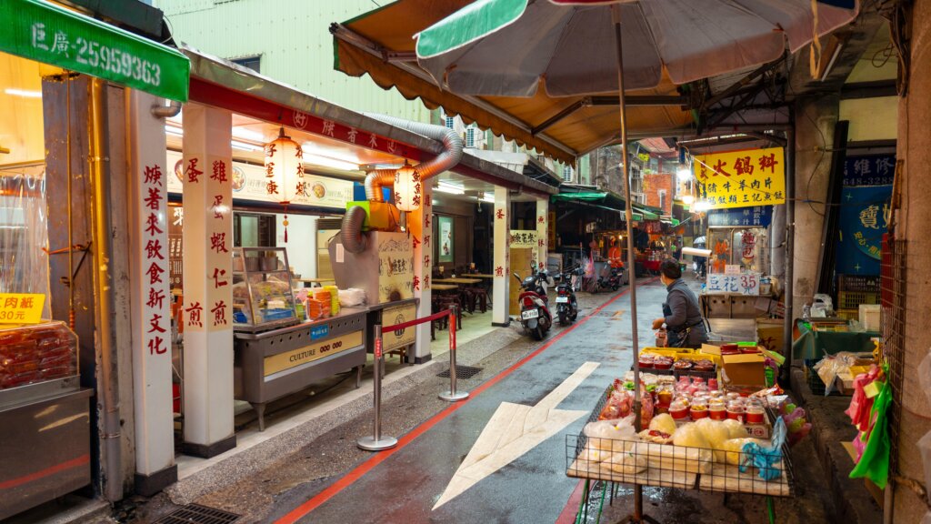Street market scene with food stalls, a vendor, and scooters under umbrellas in an Asian urban setting at dusk.
