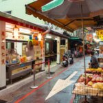 Street market scene with food stalls, a vendor, and scooters under umbrellas in an Asian urban setting at dusk.