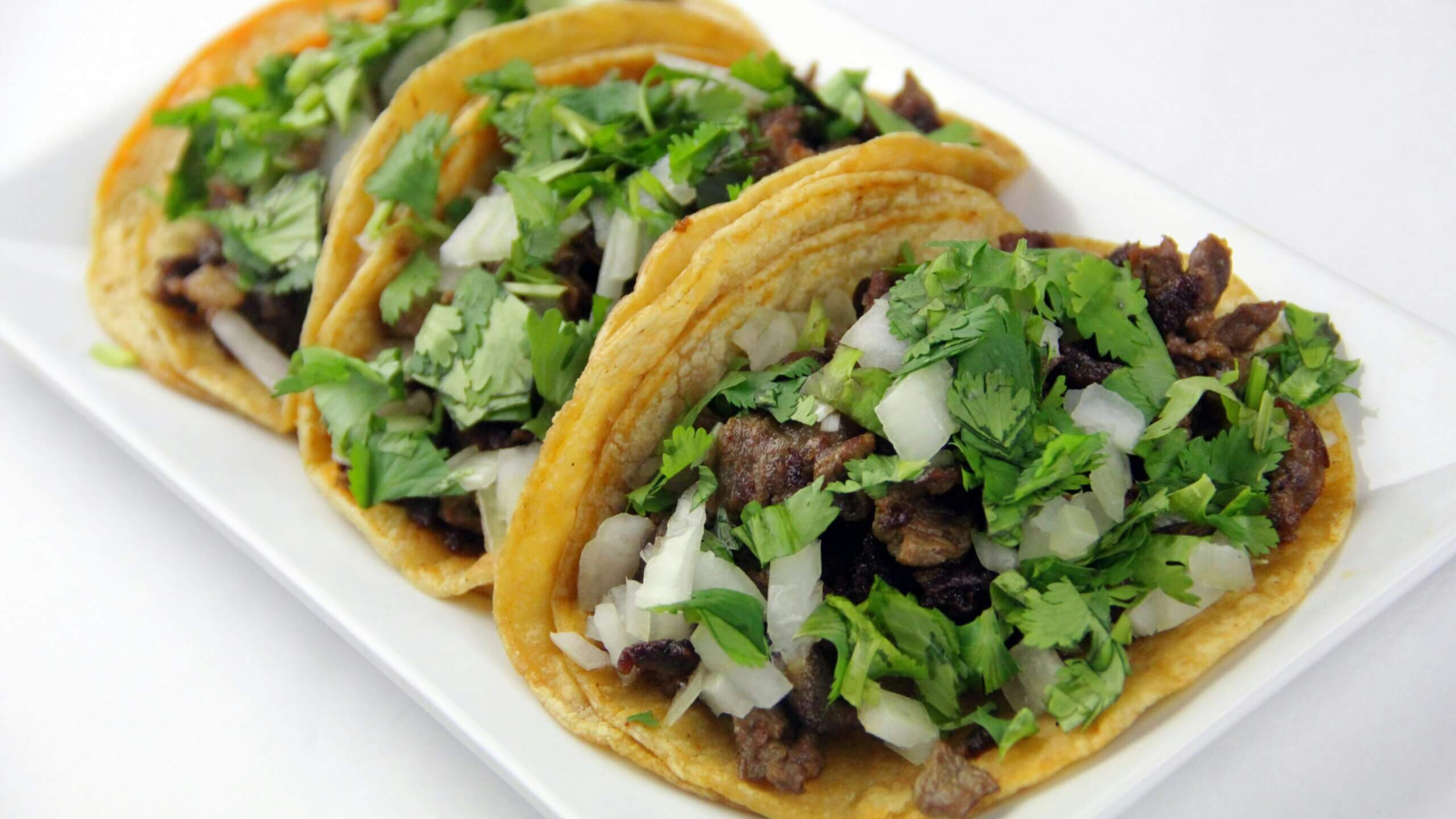 Three tacos with grilled meat, chopped onions, and cilantro on a white plate
