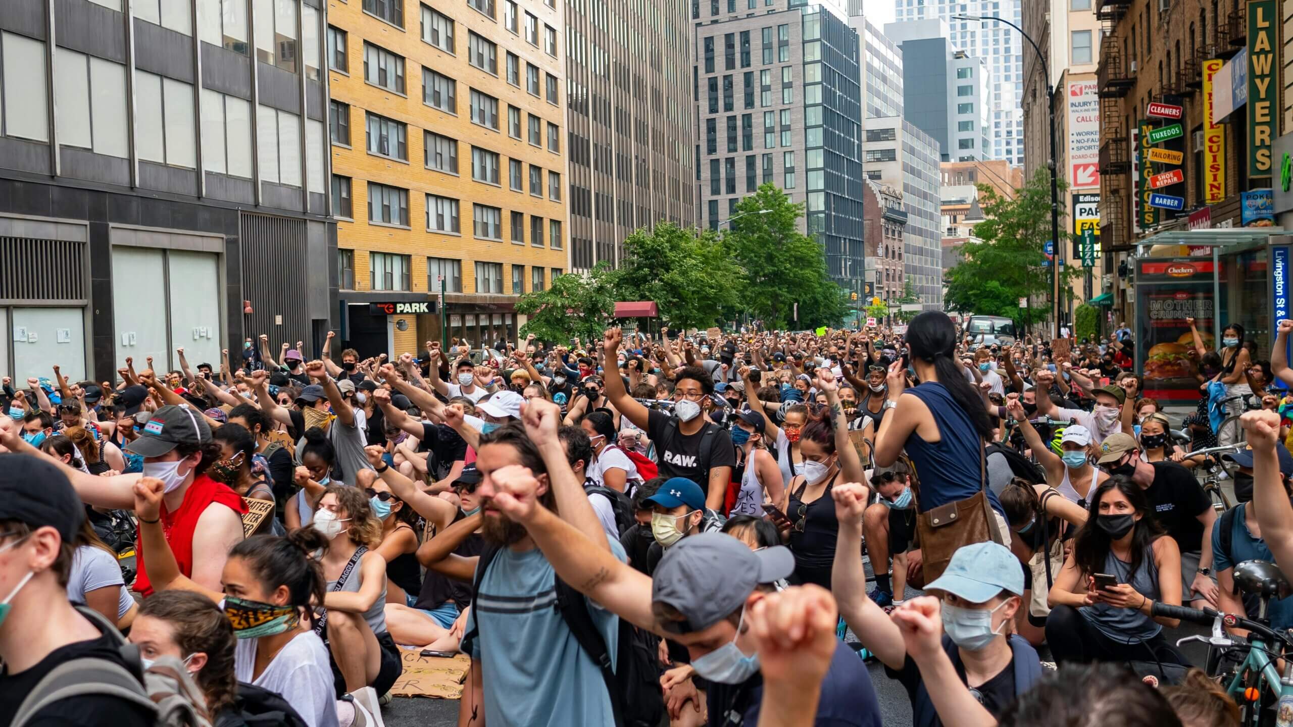 Large crowd of masked protesters sitting and raising fists in a city street during a demonstration