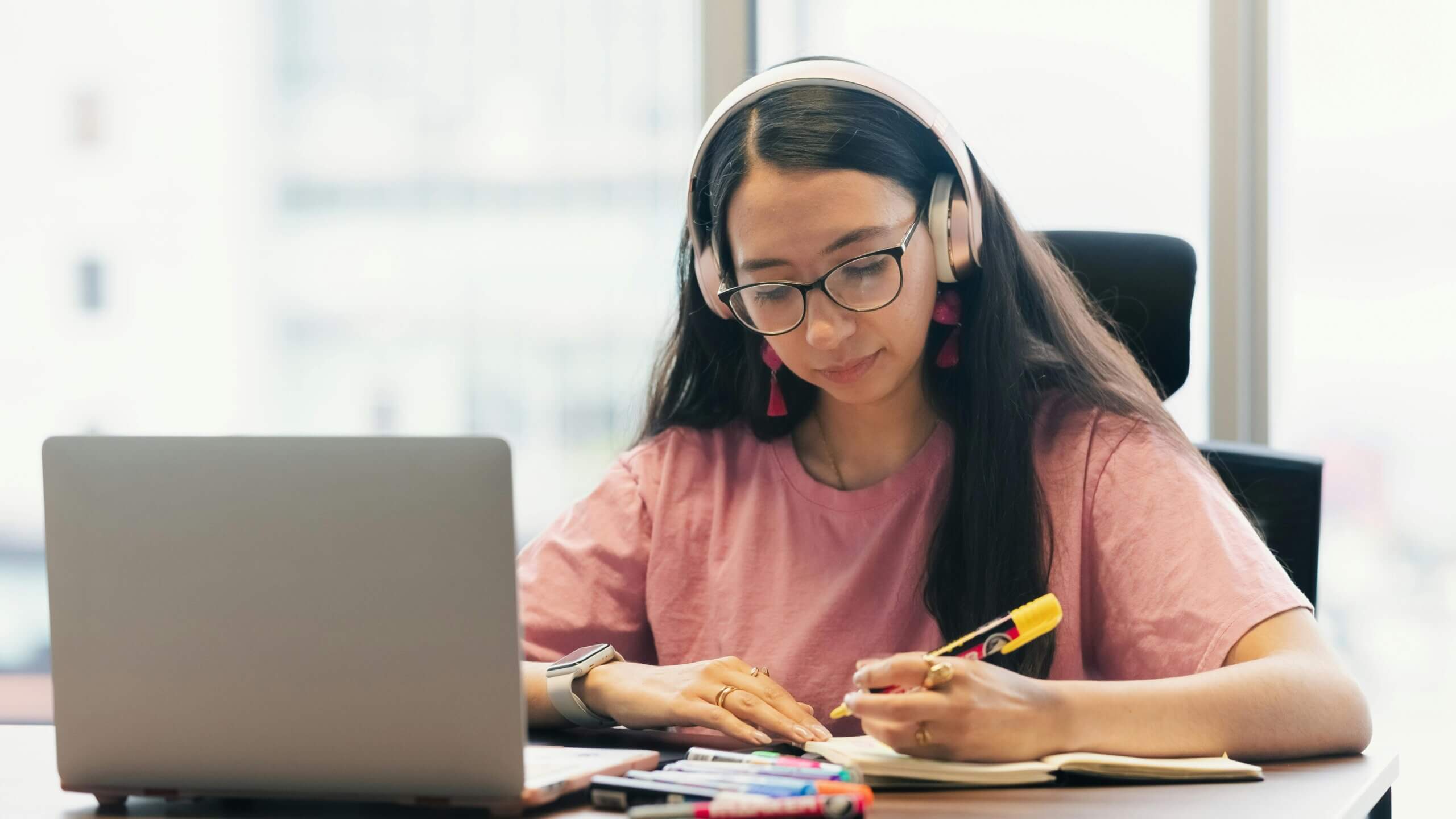 Young woman wearing glasses and headphones writing in a notebook with a laptop and markers on the desk