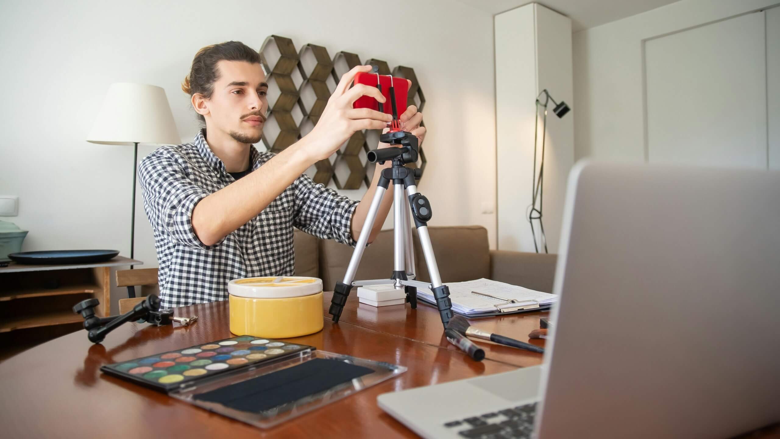 Man adjusting smartphone on tripod at table with makeup palette and laptop in home setting