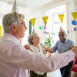 Four elderly people wearing party hats holding hands and dancing at a 60th birthday celebration with decorations.