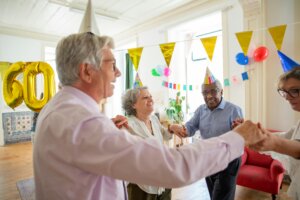 Four elderly people wearing party hats holding hands and dancing at a 60th birthday celebration with decorations.