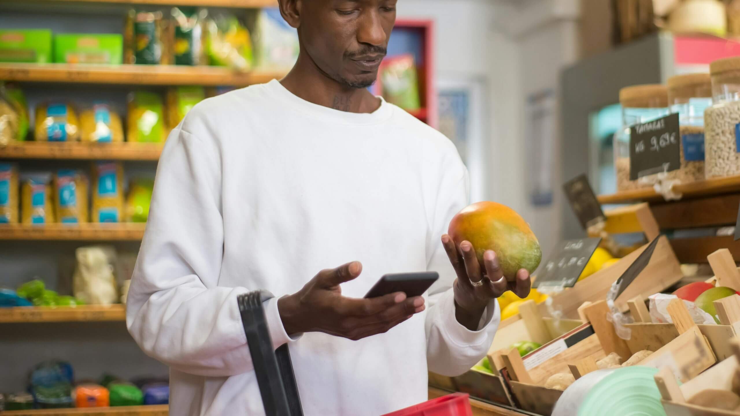 Man in white sweatshirt holding a mango and using a smartphone in a grocery store produce section