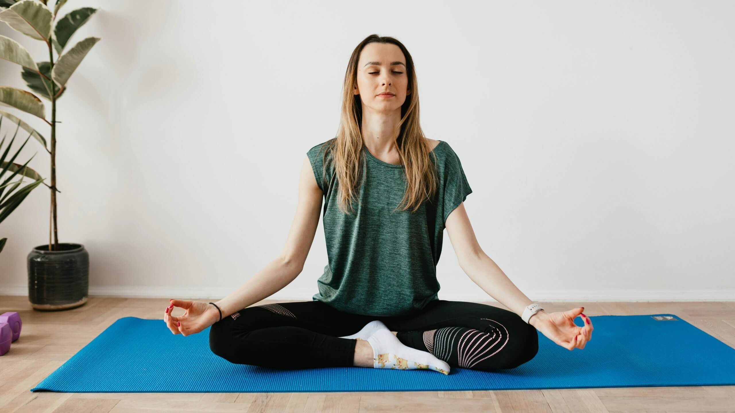 Woman meditating cross-legged on a blue yoga mat indoors with eyes closed and hands in mudra position.