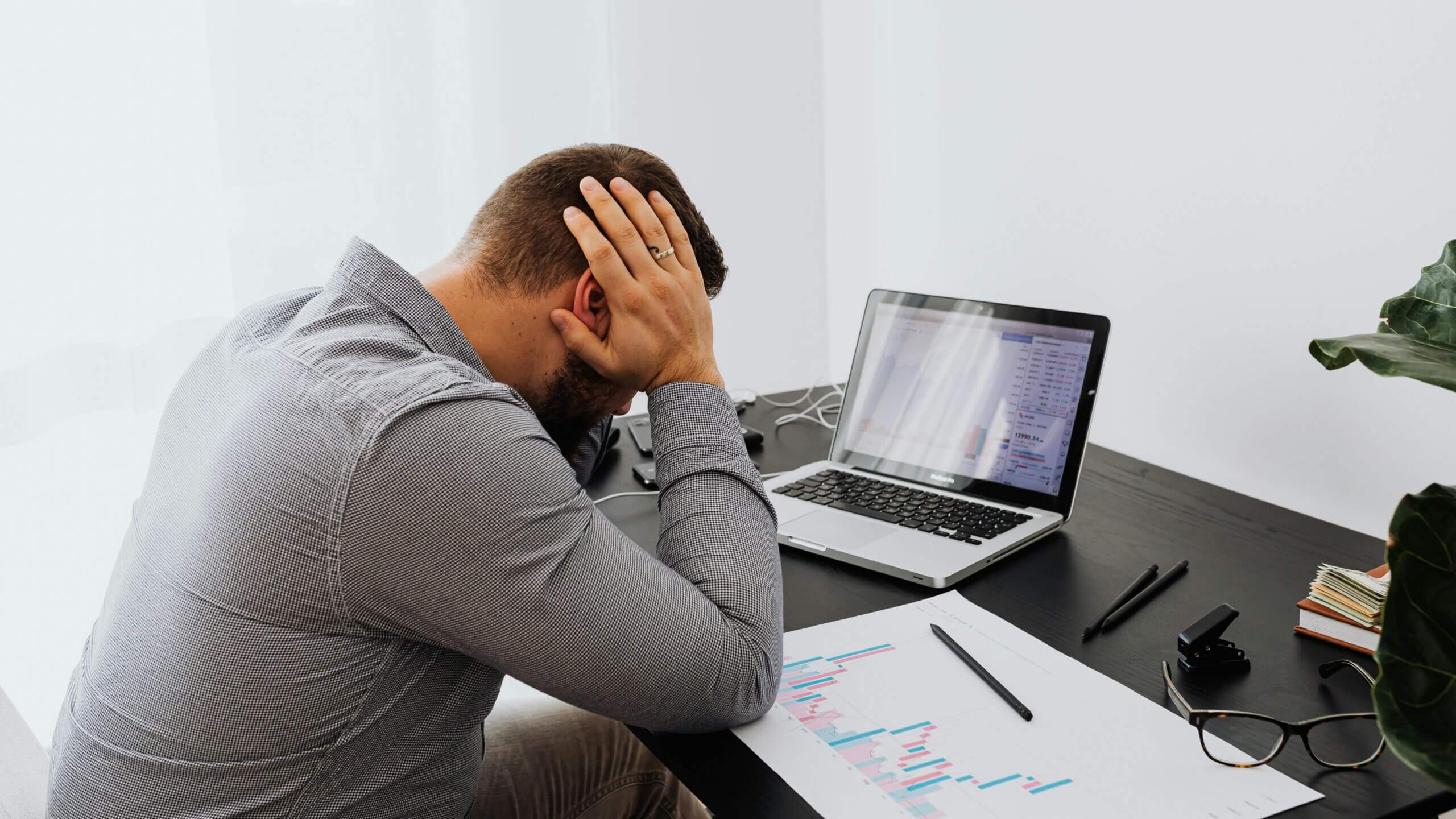 Man in gray shirt holding his head at a desk with a laptop showing financial charts and printed graphs.