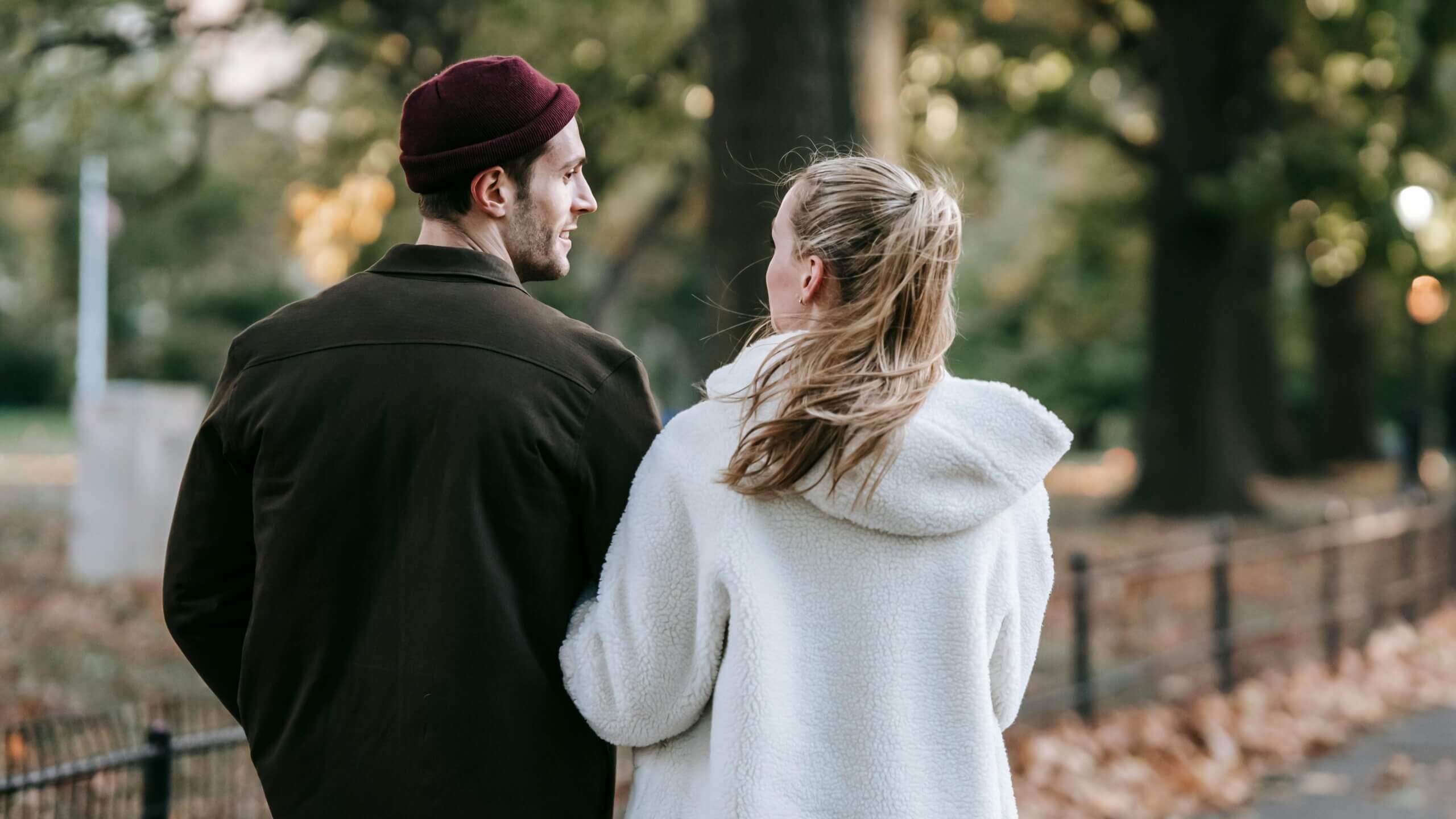 Couple walking and talking in a park during autumn, man wearing a maroon beanie and dark jacket, woman in a white fleece jacket