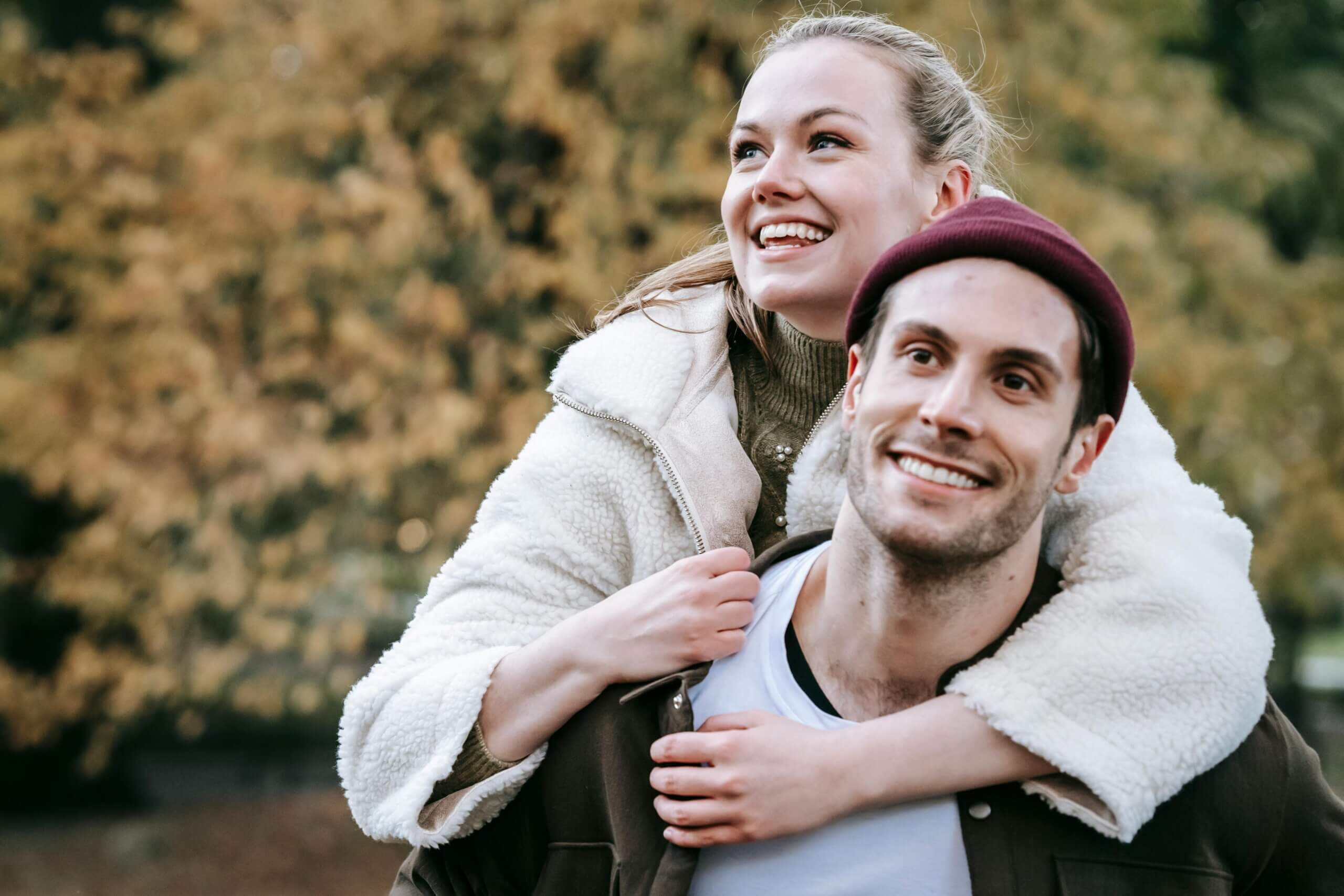 Smiling woman in white fleece jacket piggybacking on a man wearing a maroon beanie outdoors in autumn.