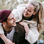 Smiling woman in white fleece jacket piggybacking on man wearing maroon beanie and brown jacket outdoors