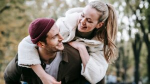Smiling woman in white fleece jacket piggybacking on man wearing maroon beanie and brown jacket outdoors
