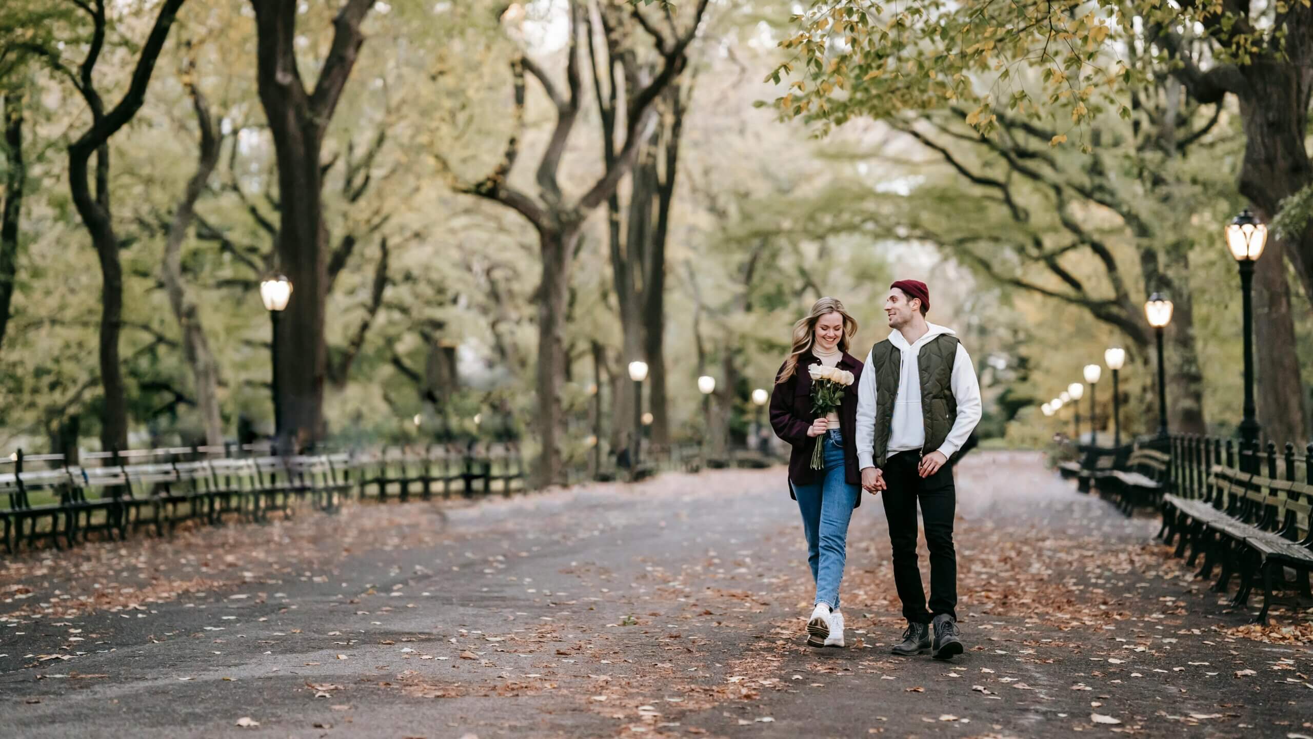Couple holding hands walking in a tree-lined park path with fallen autumn leaves and street lamps on either side