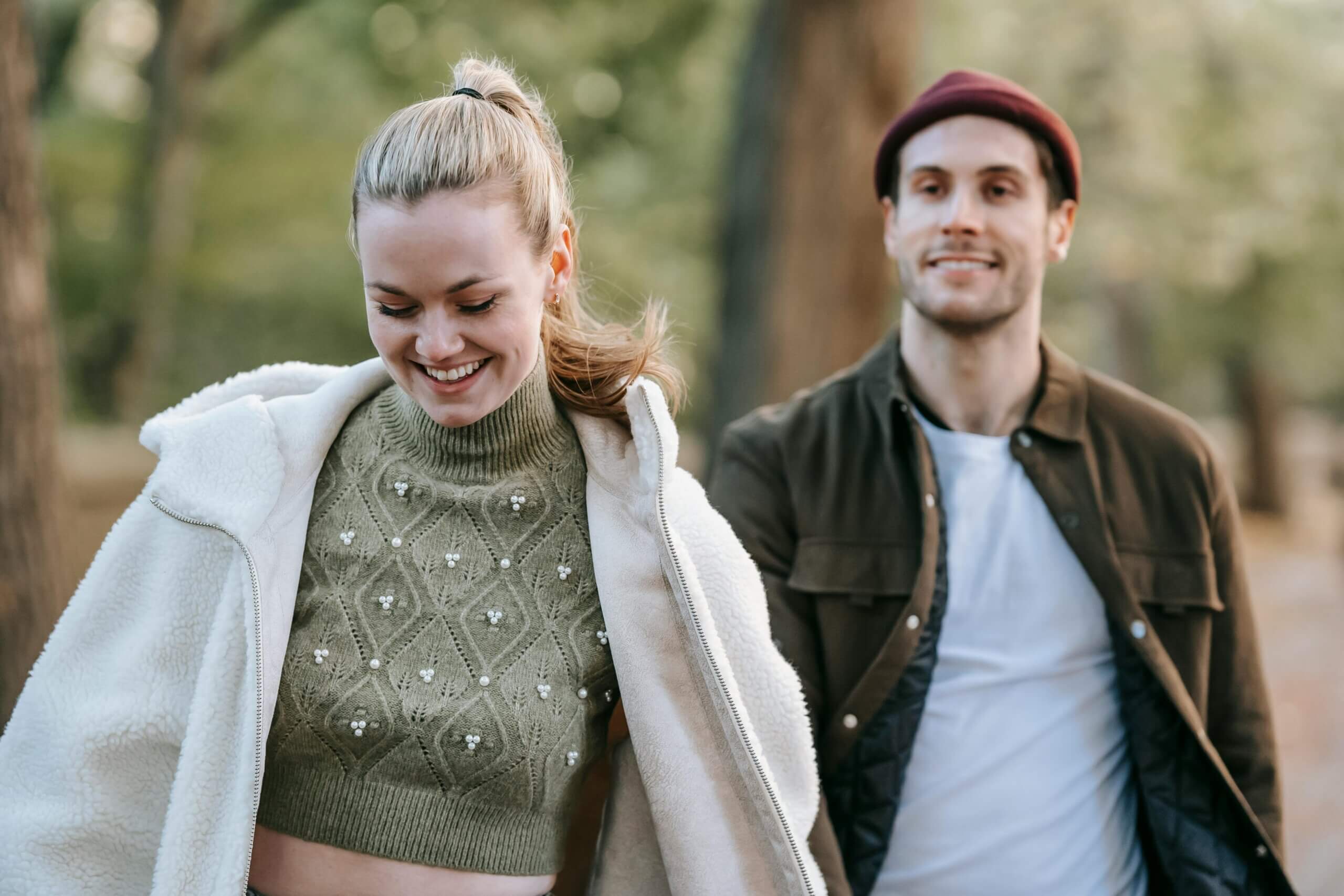 Smiling woman in a green knit sweater and white jacket walks outdoors with a man in a brown jacket and red beanie.