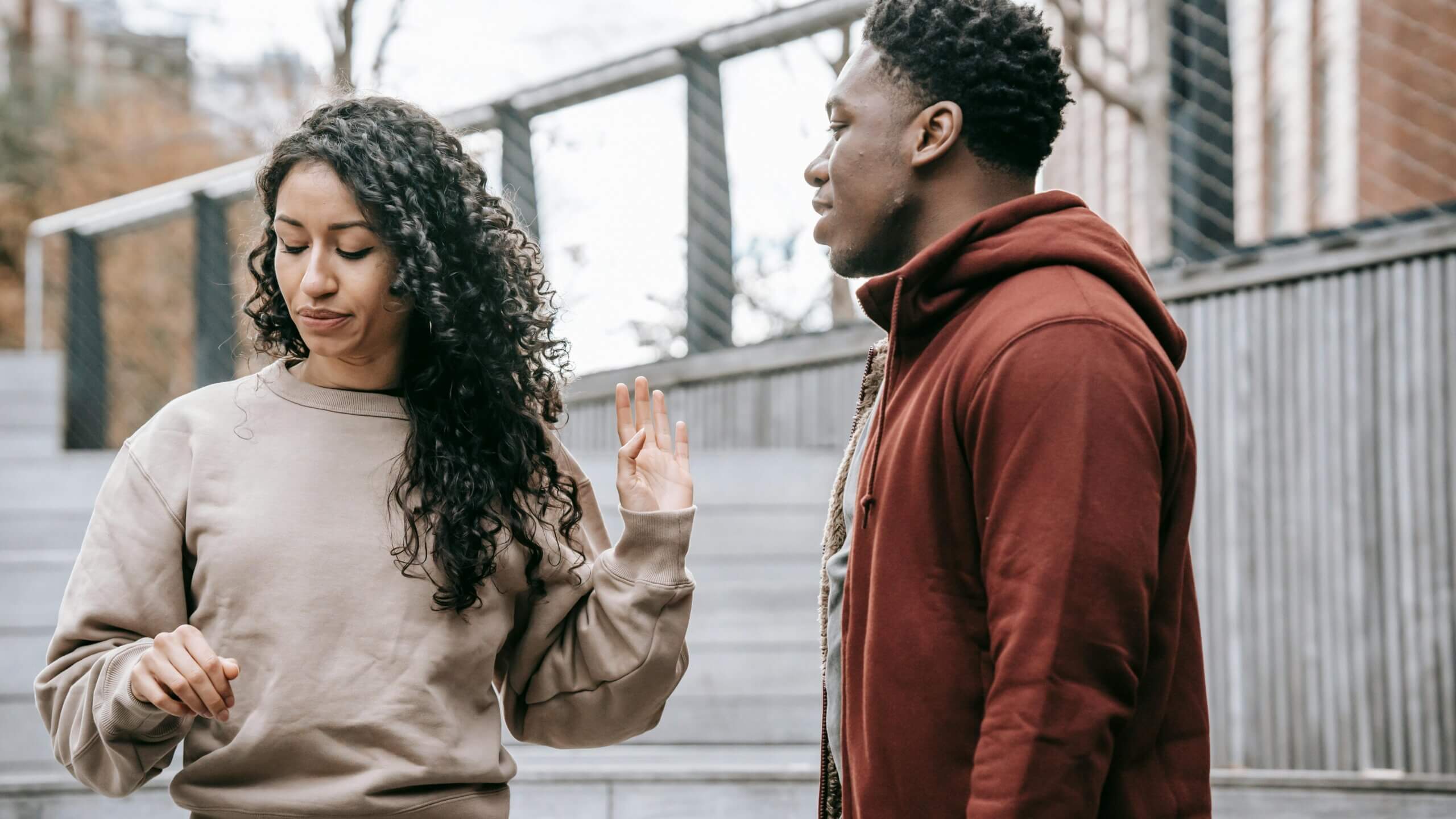 Woman with curly hair in beige sweatshirt turning away from man in red hoodie during outdoor conversation