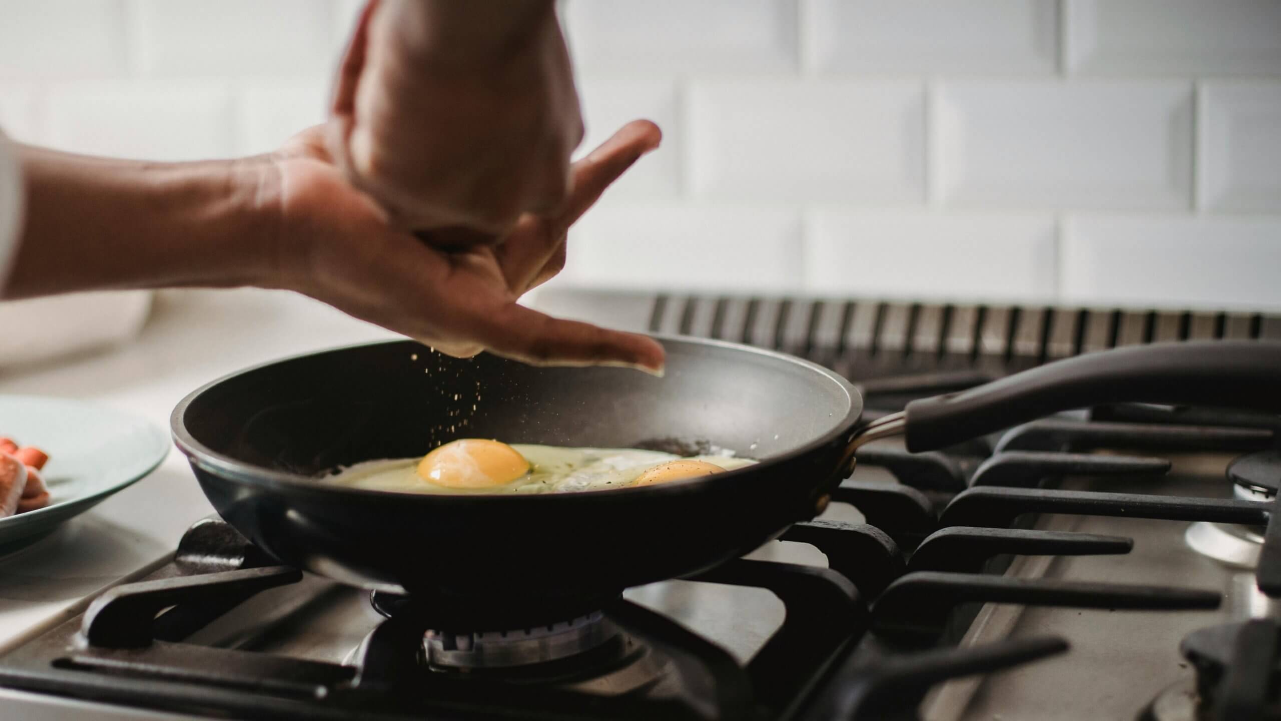 Hand sprinkling seasoning onto two eggs frying in a black pan on a gas stove.
