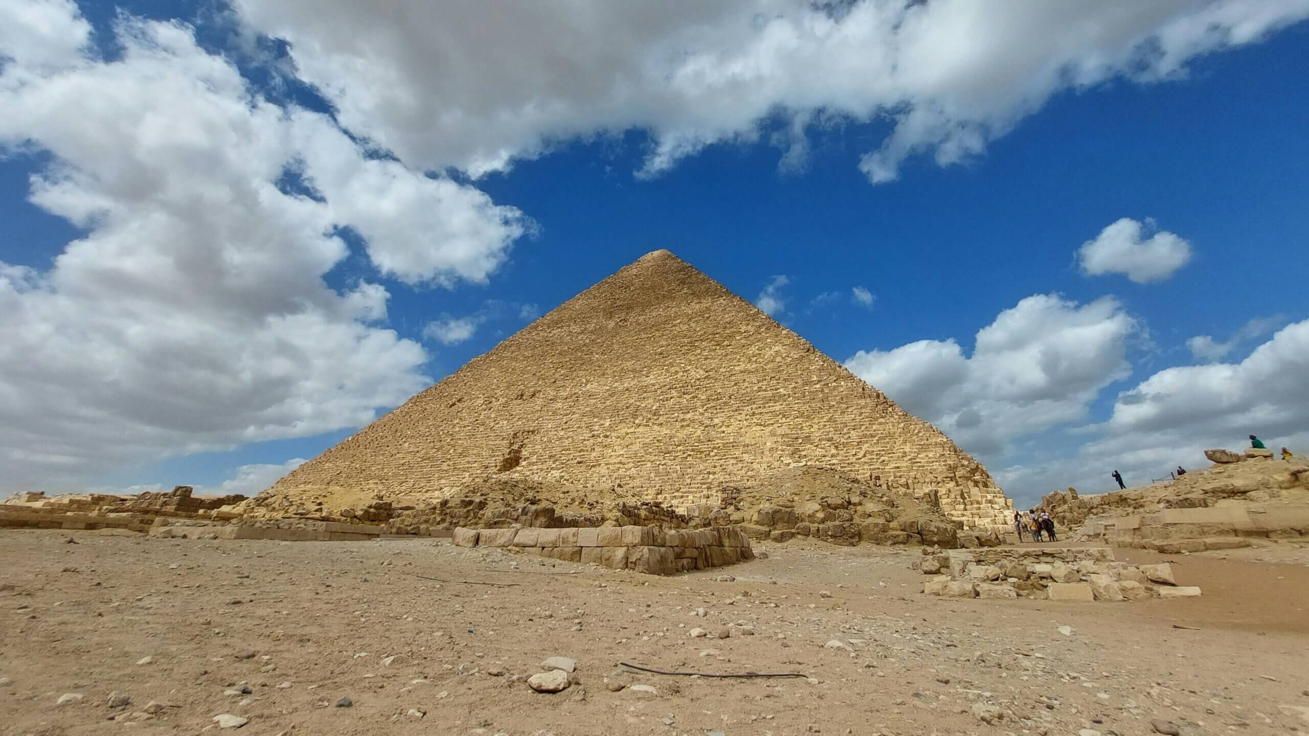 The Great Pyramid of Giza under a partly cloudy blue sky with scattered tourists nearby.