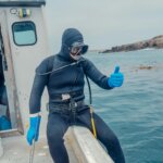 Scuba diver in black wetsuit and blue gloves giving thumbs up while sitting on a boat near rocky coastline