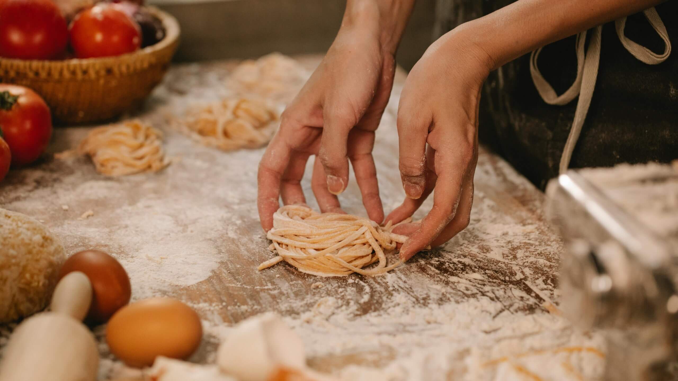 Hands shaping fresh pasta nests on a floured wooden surface with tomatoes, eggs, and a pasta machine nearby.