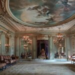 Ornate throne room with gilded moldings, chandeliers, purple drapery, and a ceiling fresco of mythological figures.