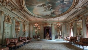 Ornate throne room with gilded moldings, chandeliers, purple drapery, and a ceiling fresco of mythological figures.