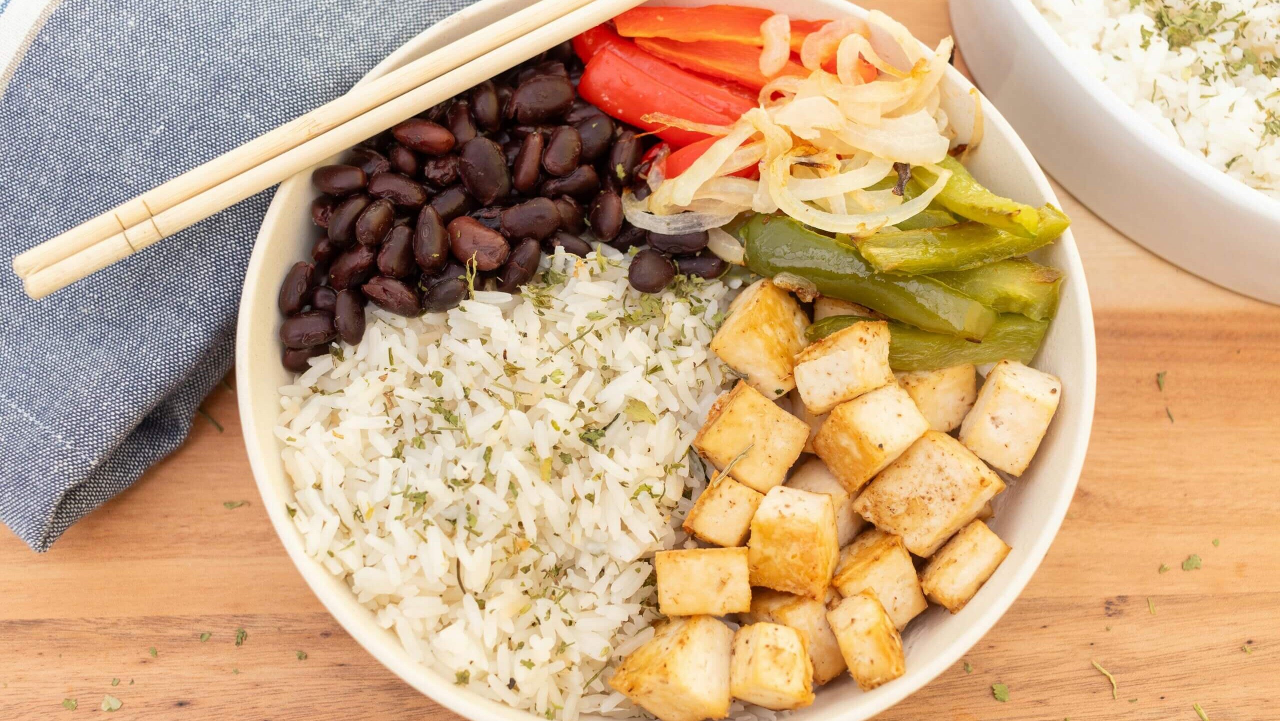 Bowl with white rice, black beans, sautéed tofu, grilled bell peppers, and onions with chopsticks on a napkin.