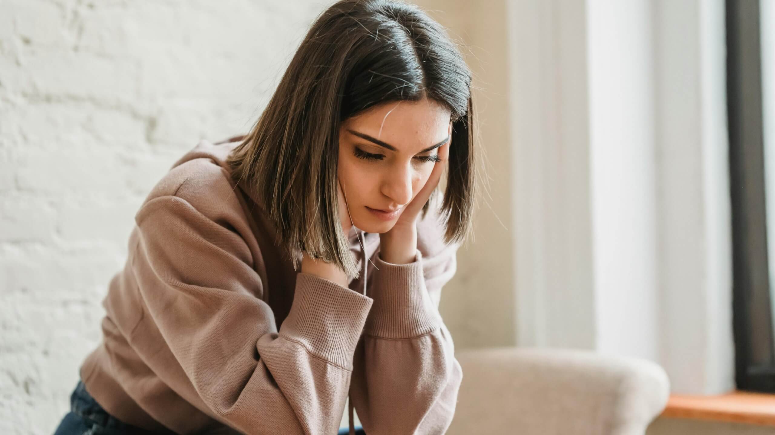 Young woman in a mauve sweatshirt looking down with hands on her neck near a window and white brick wall background