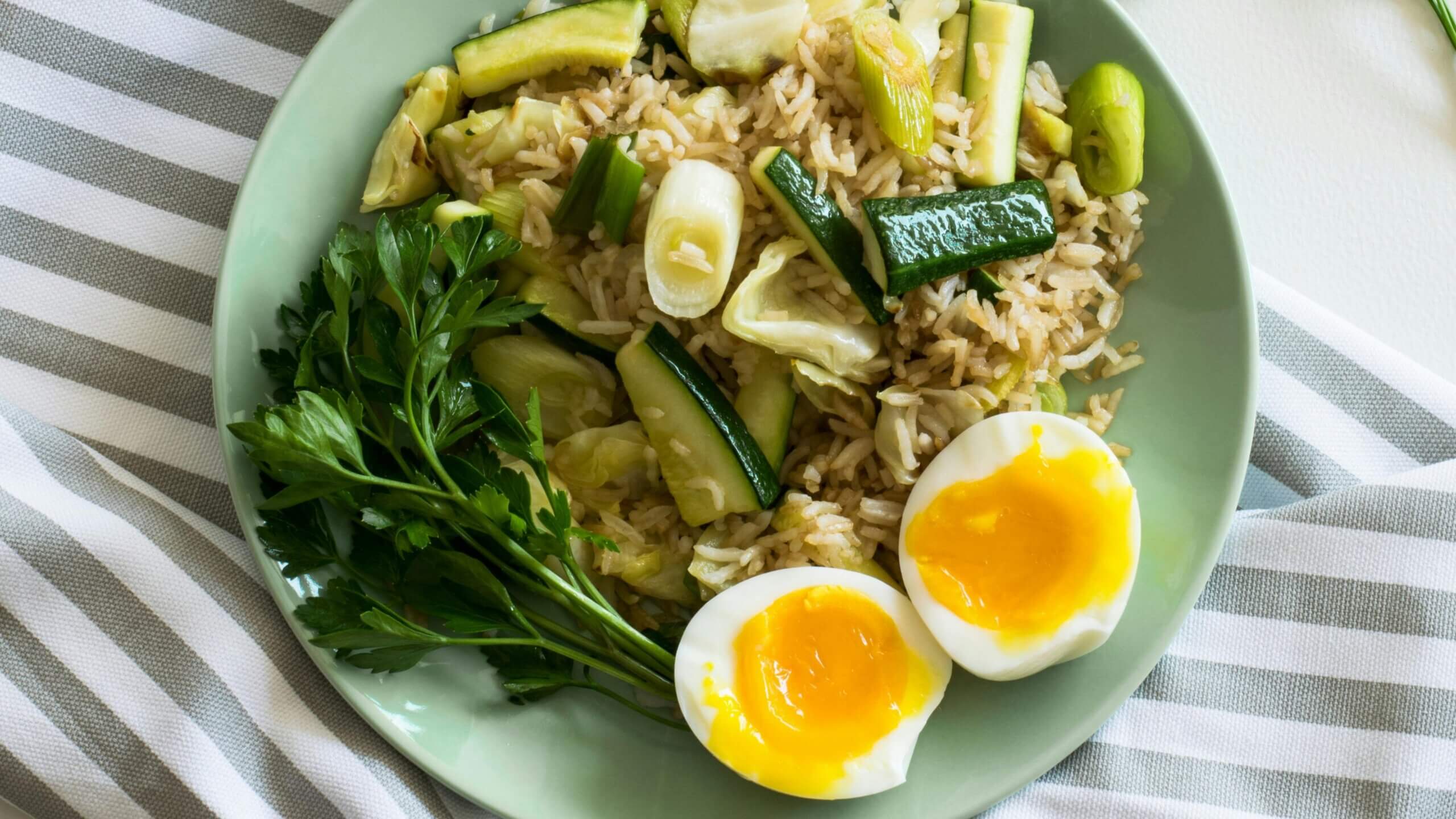 Plate with rice, cooked zucchini, soft-boiled eggs, and fresh parsley on a striped cloth background