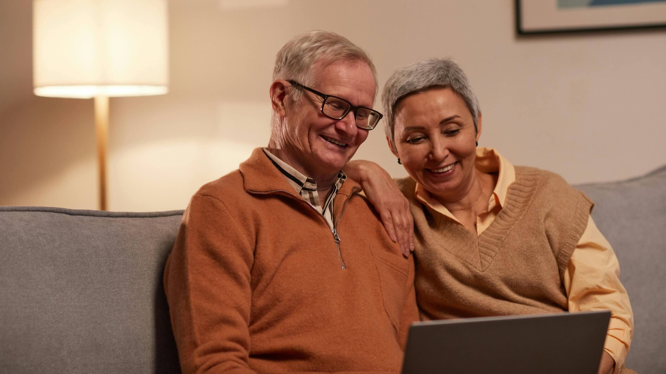 Smiling elderly couple sitting on a couch looking at a laptop together in a warmly lit room.