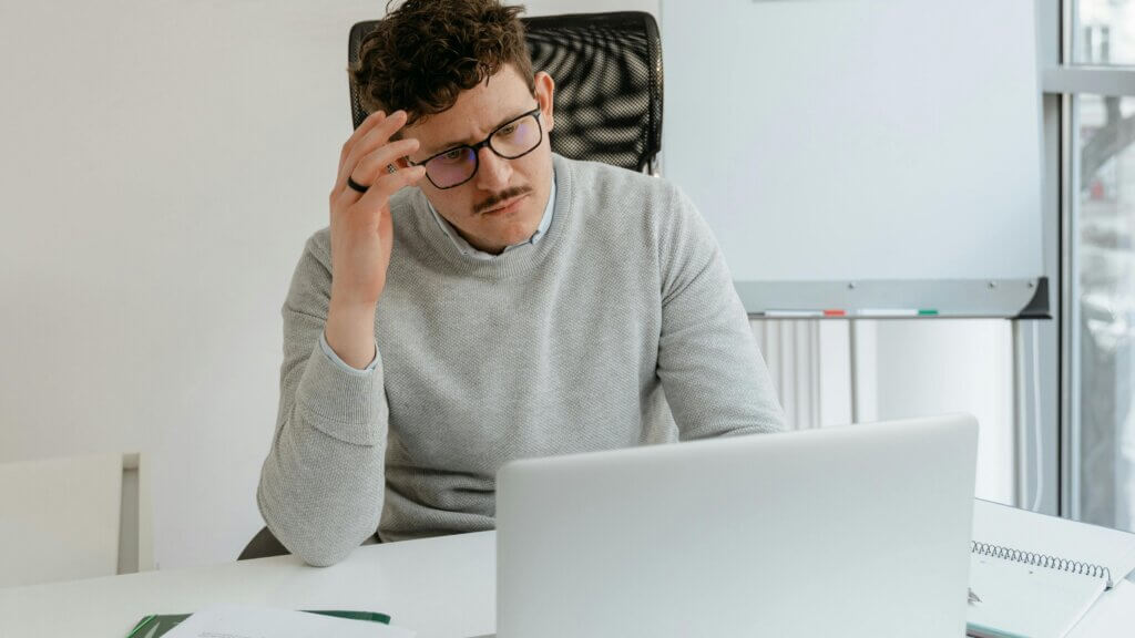 Man with glasses and mustache wearing a gray sweater, looking confused at a laptop in an office setting