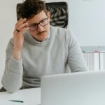 Man with glasses and mustache wearing a gray sweater, looking confused at a laptop in an office setting