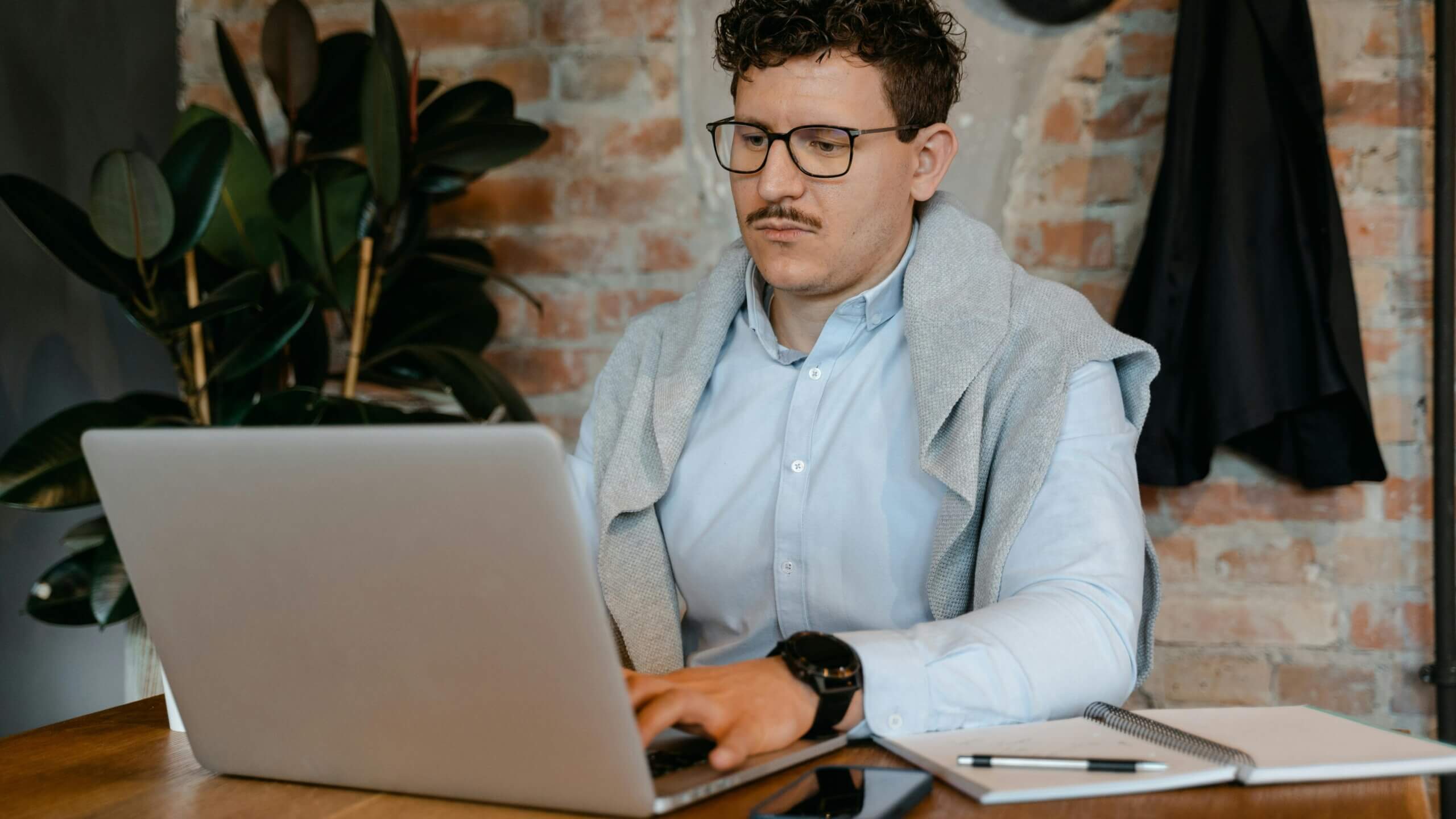 Man with glasses and mustache working on a laptop at a wooden desk with a notebook and phone nearby
