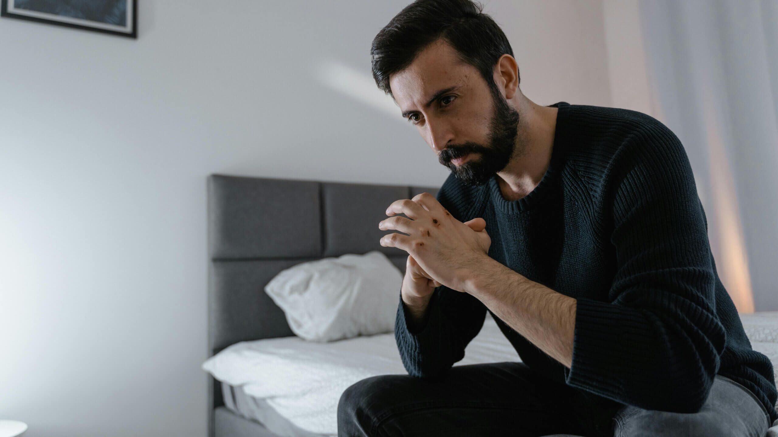 Man with beard and black sweater sitting on bed, looking thoughtful with hands clasped and visible scars on knuckles.