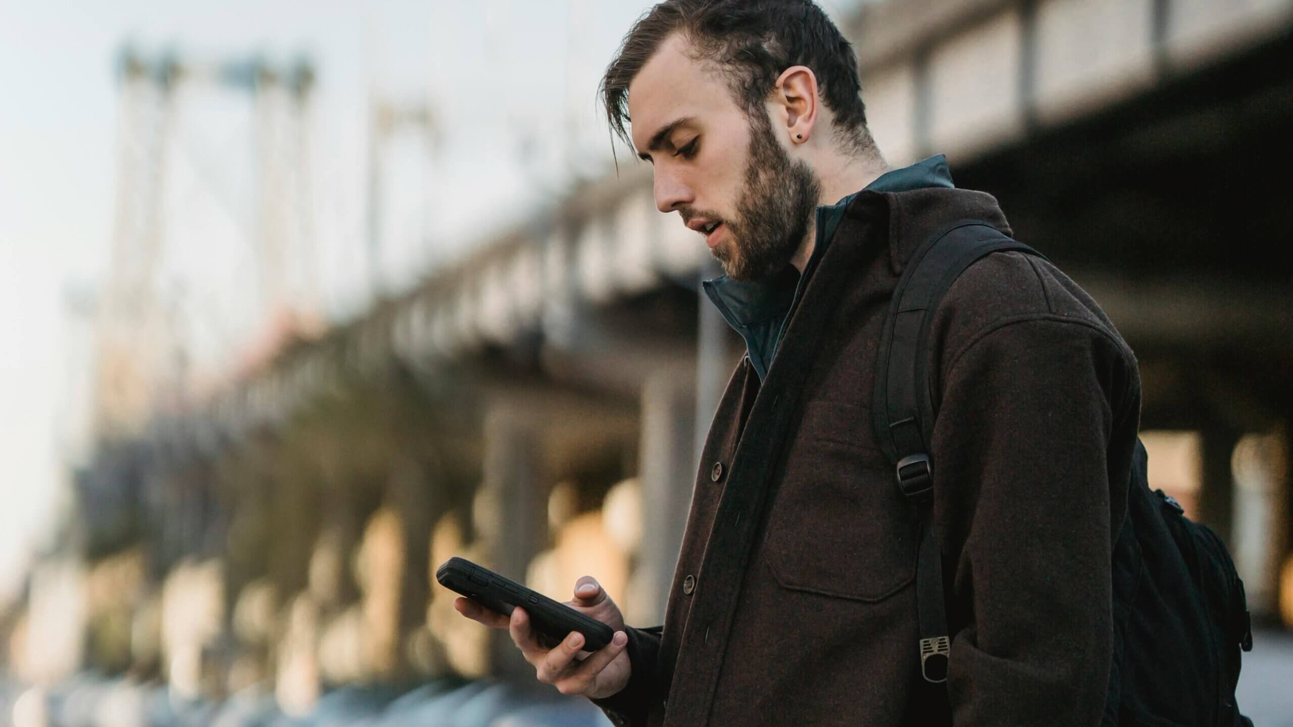 Young man with beard and backpack looking at smartphone outdoors near a bridge