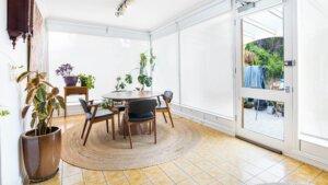 Dining area with round wooden table, four chairs, indoor plants, and large windows with white blinds.