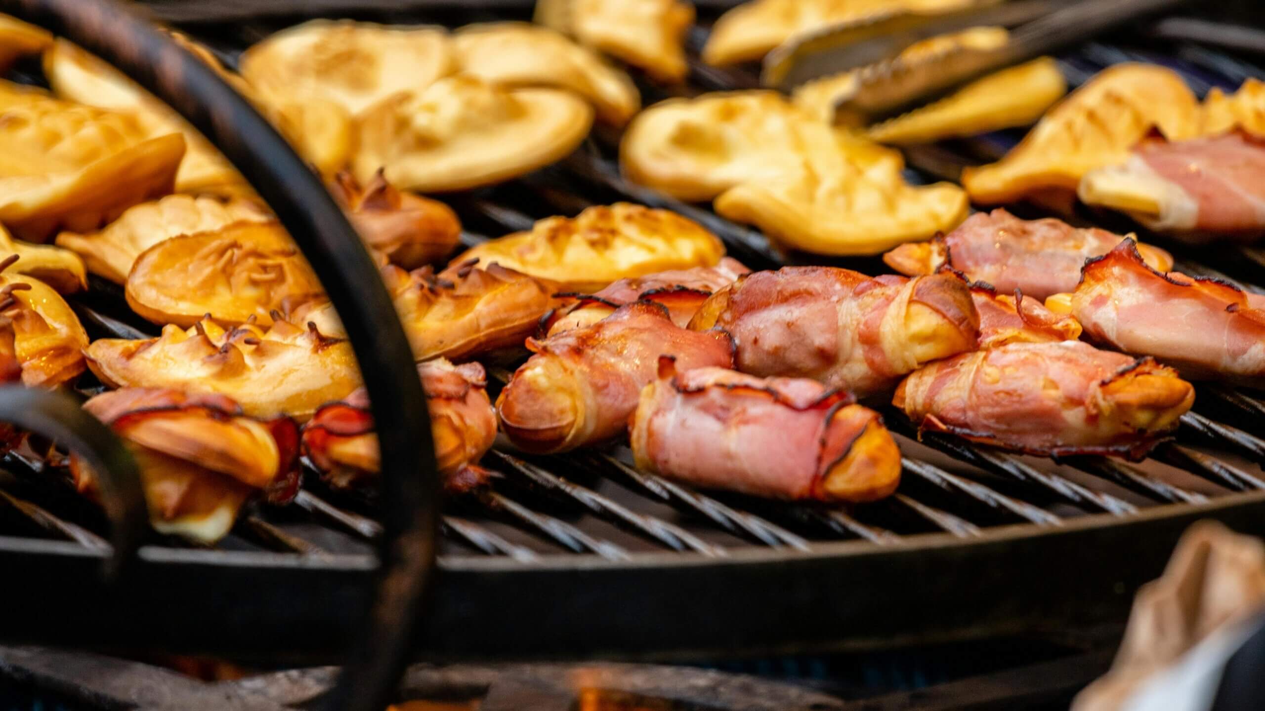 Bacon-wrapped sausages and grilled pastries cooking on a barbecue grill.