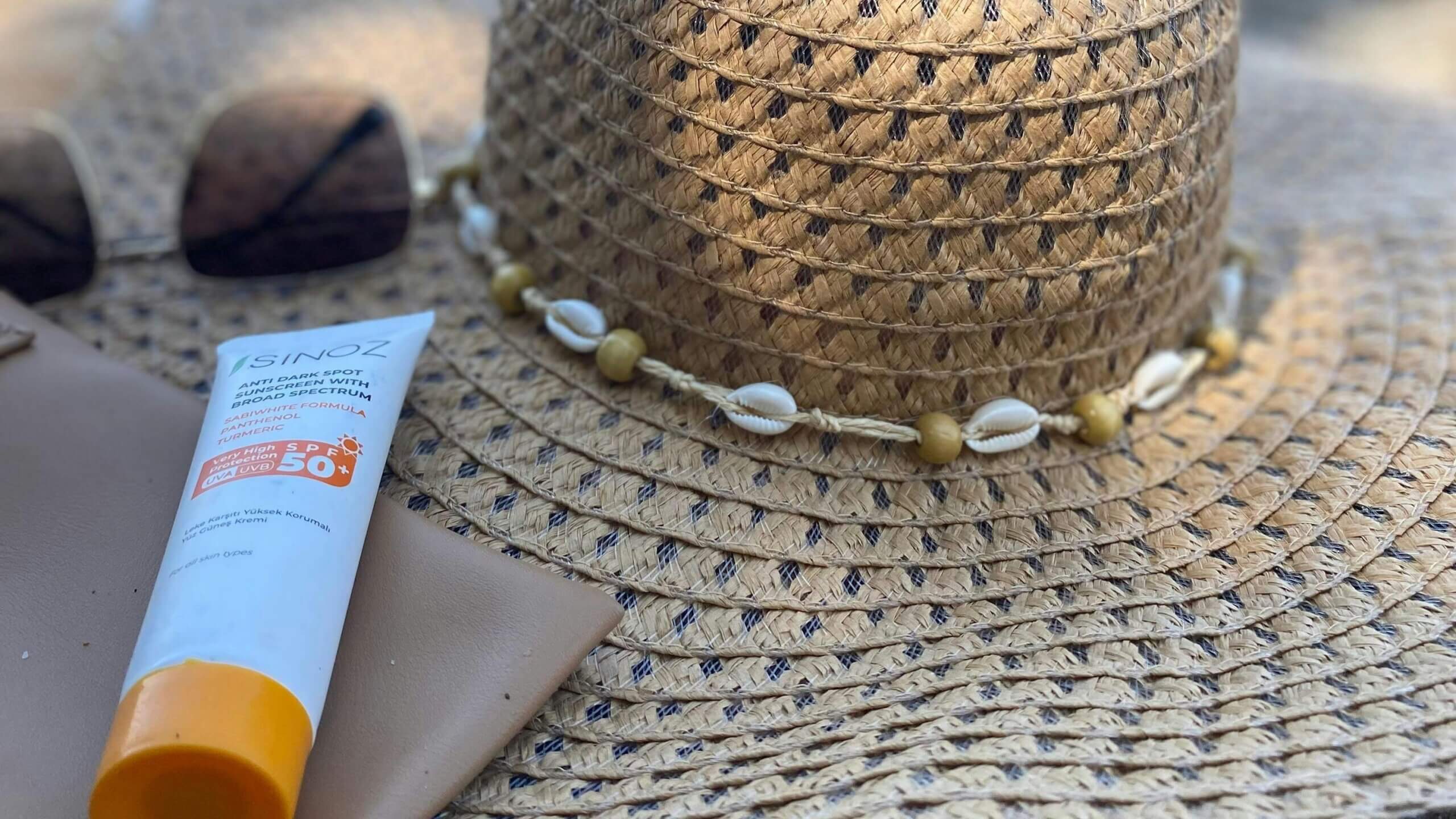 Sinoz SPF 50+ anti dark spot sunscreen tube next to a straw hat with shell and bead decoration and sunglasses