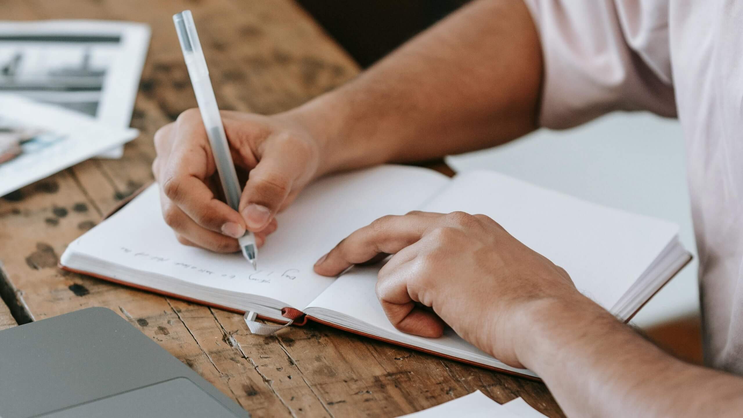 Person writing in a notebook with a pen on a wooden table next to a laptop and papers