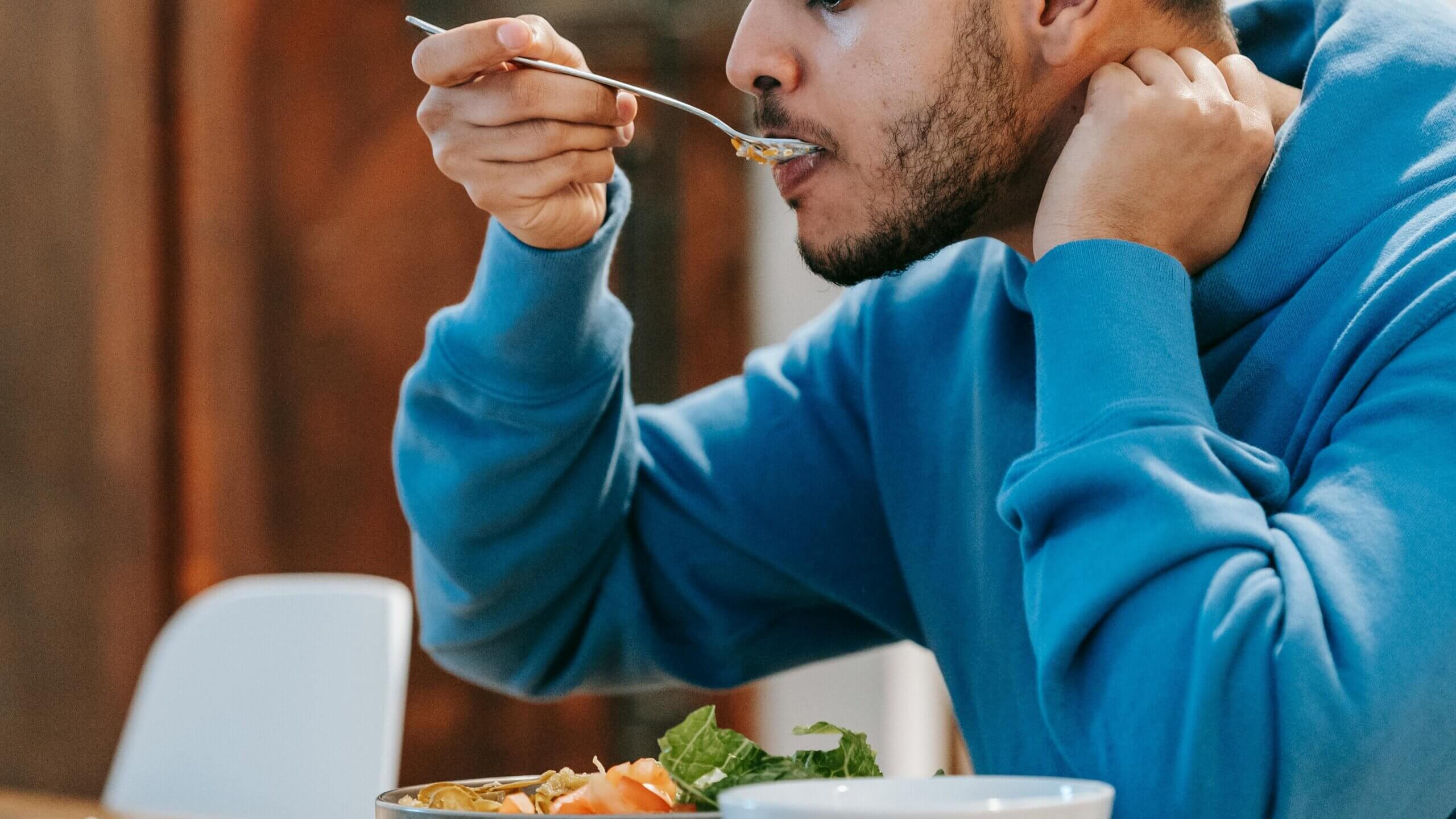 Man in blue hoodie eating salad with a spoon at a wooden table