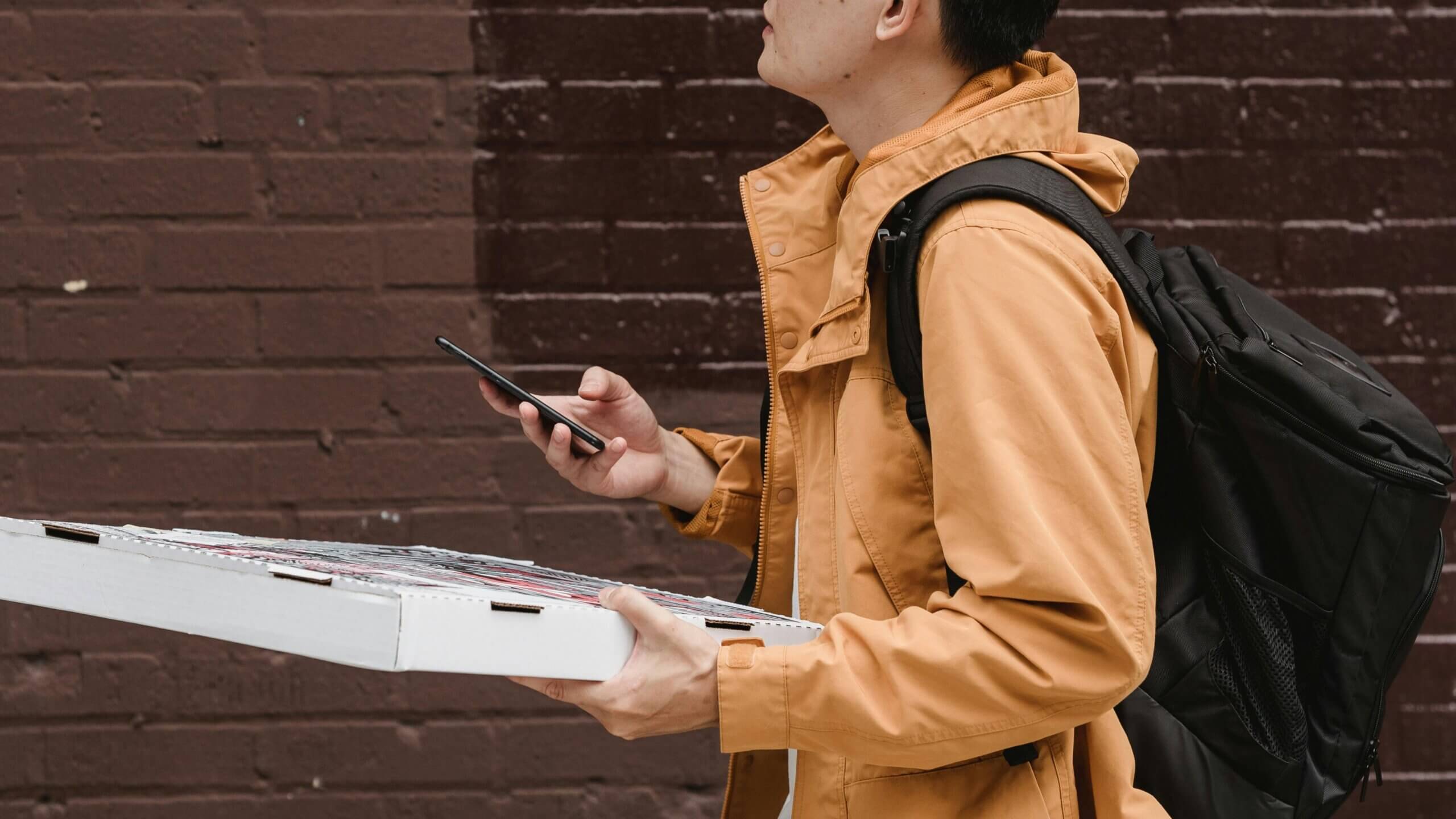 Person in tan jacket holding pizza boxes and using a smartphone against a brown brick wall background