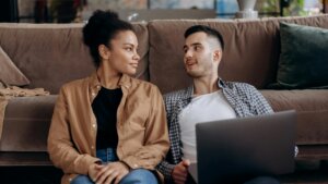 Young man and woman sitting on the floor, talking with a laptop in a cozy living room.