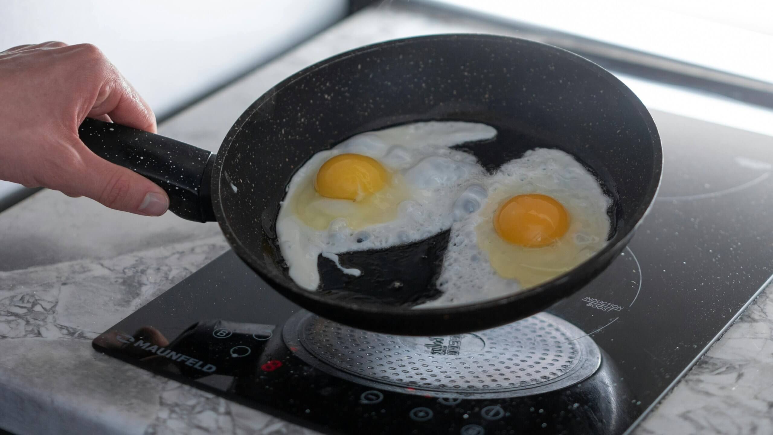 Hand holding a black speckled frying pan with two eggs cooking on a Maunfeld induction stove.