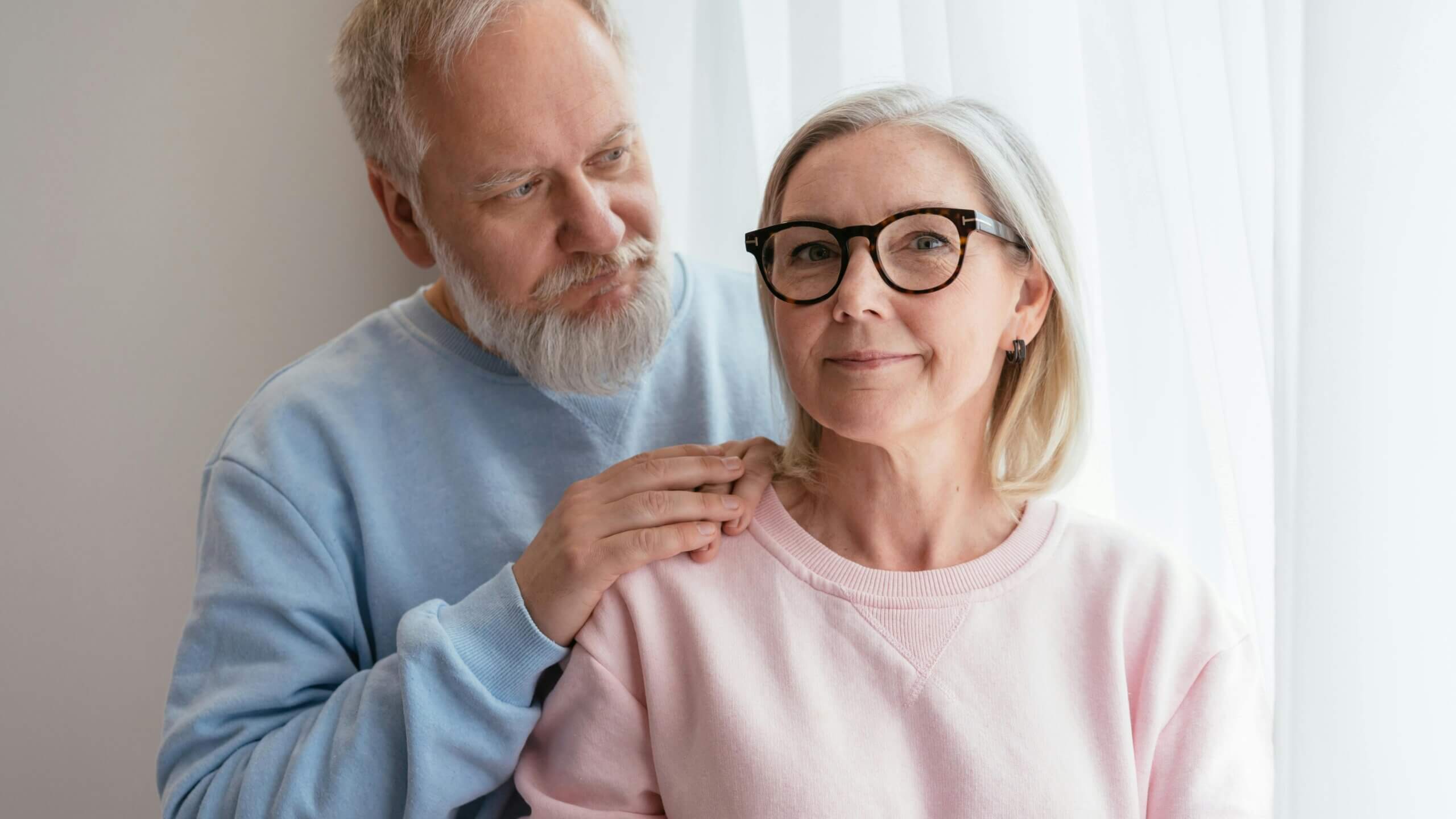 Older man in blue sweater comforting older woman in pink sweater and glasses by touching her shoulder