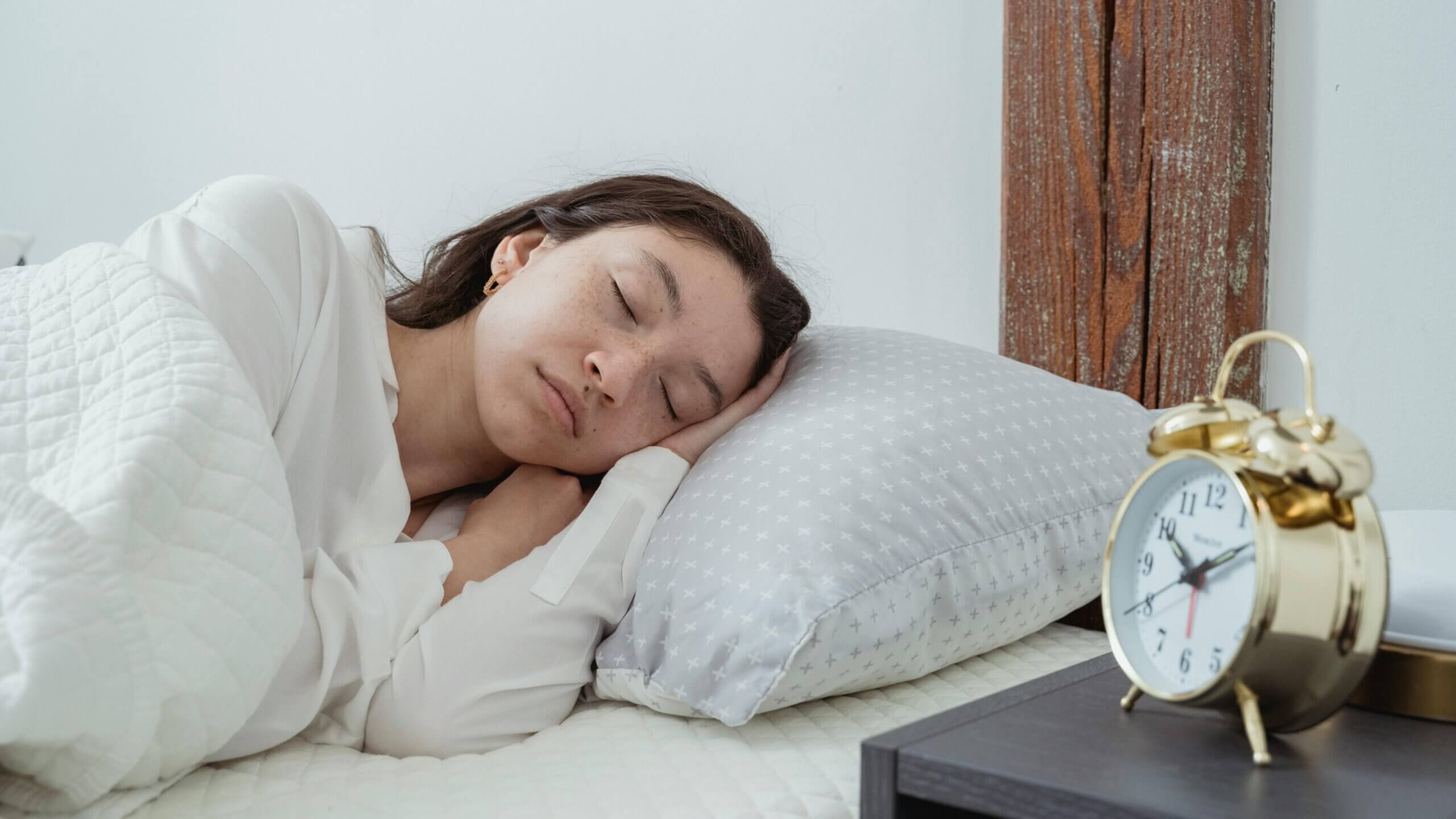 Woman sleeping on bed with white quilt and gray pillow next to gold alarm clock showing 7:10