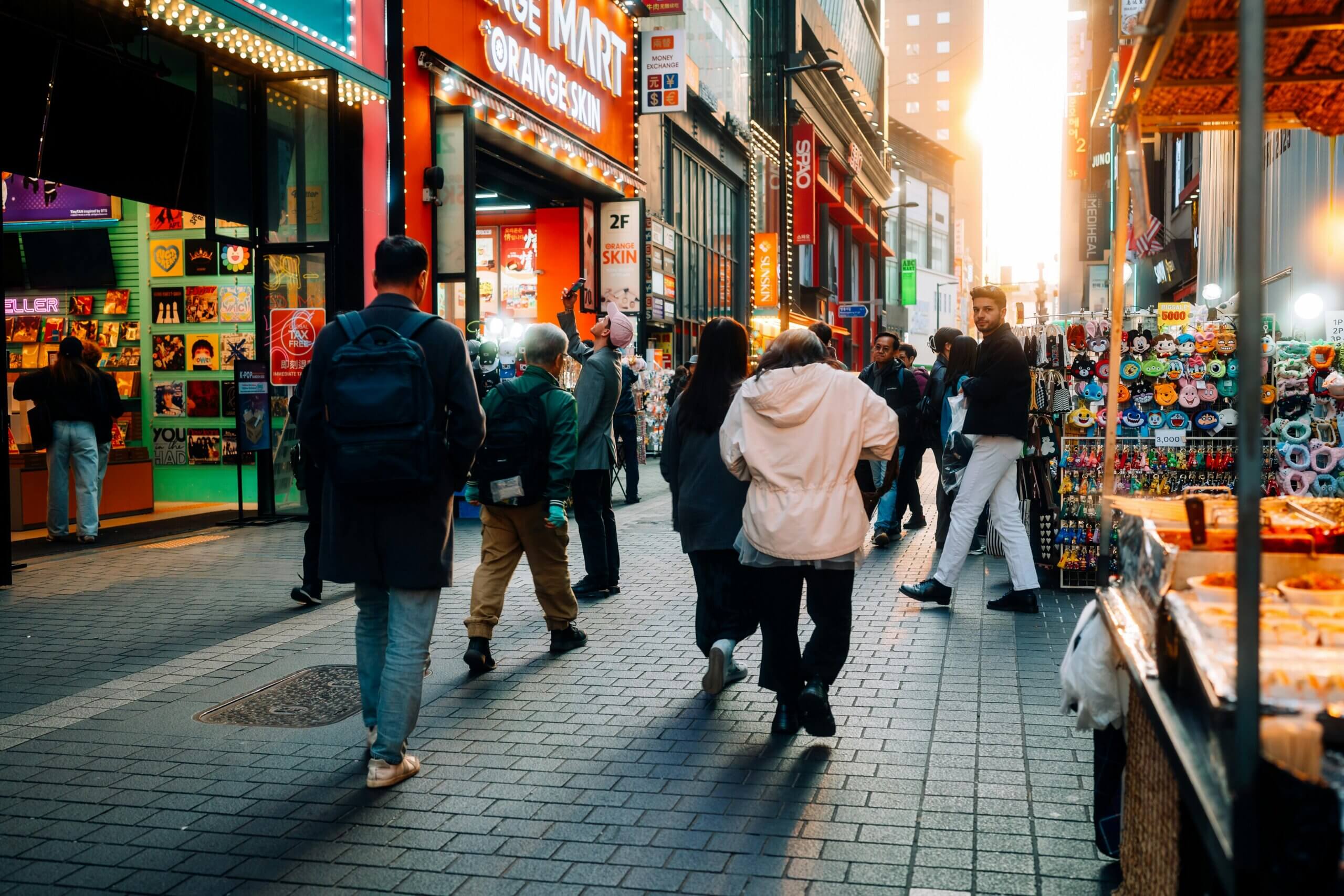 People walking and shopping on a busy street near Orange Mart and souvenir stalls at sunset.