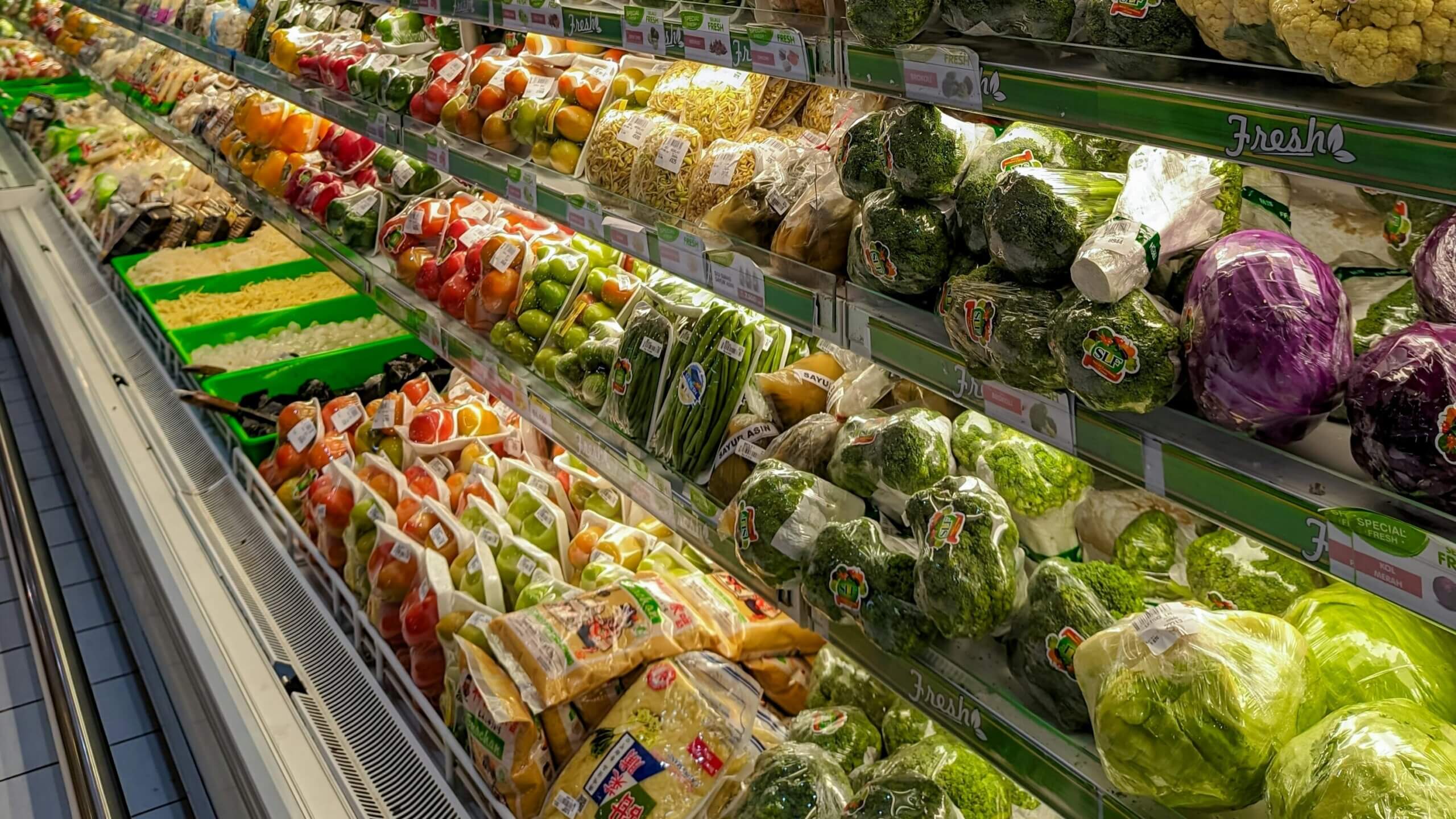 Packaged fresh vegetables including broccoli, cabbage, bell peppers, and green beans on supermarket shelves labeled "Fresh"