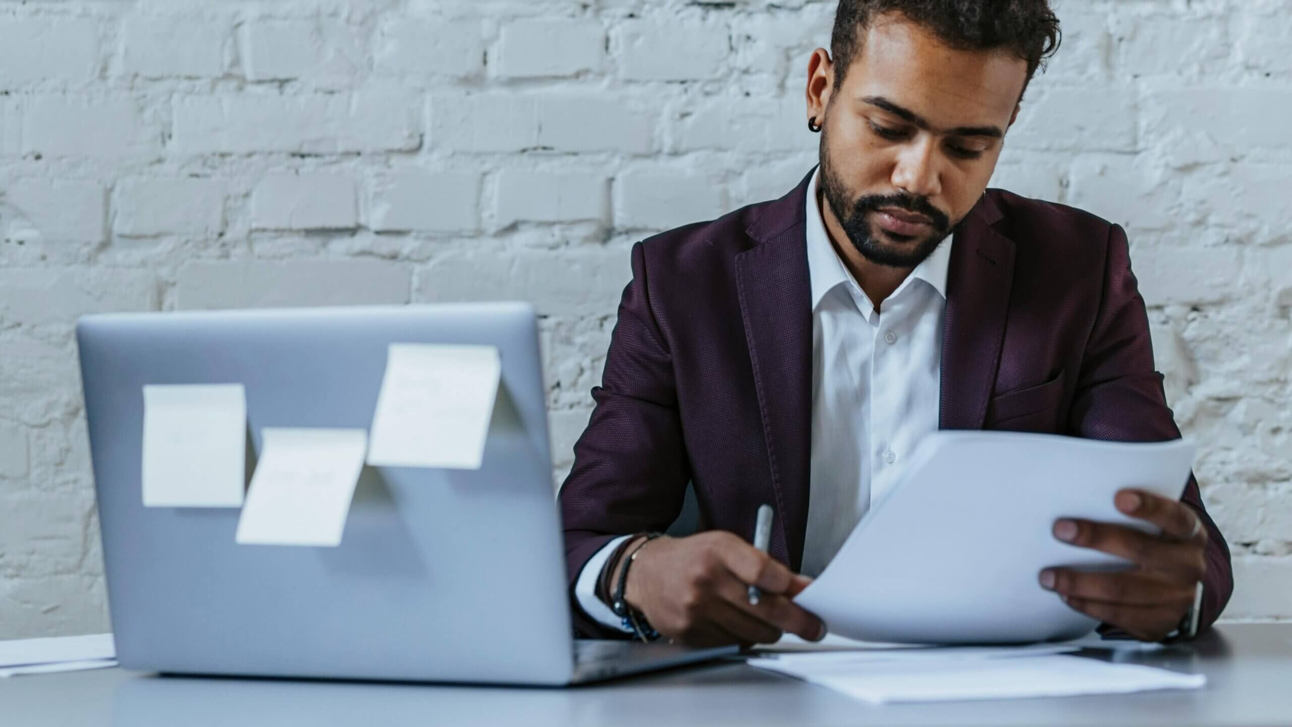 Man in a burgundy blazer reviewing documents at a desk with a laptop covered in sticky notes