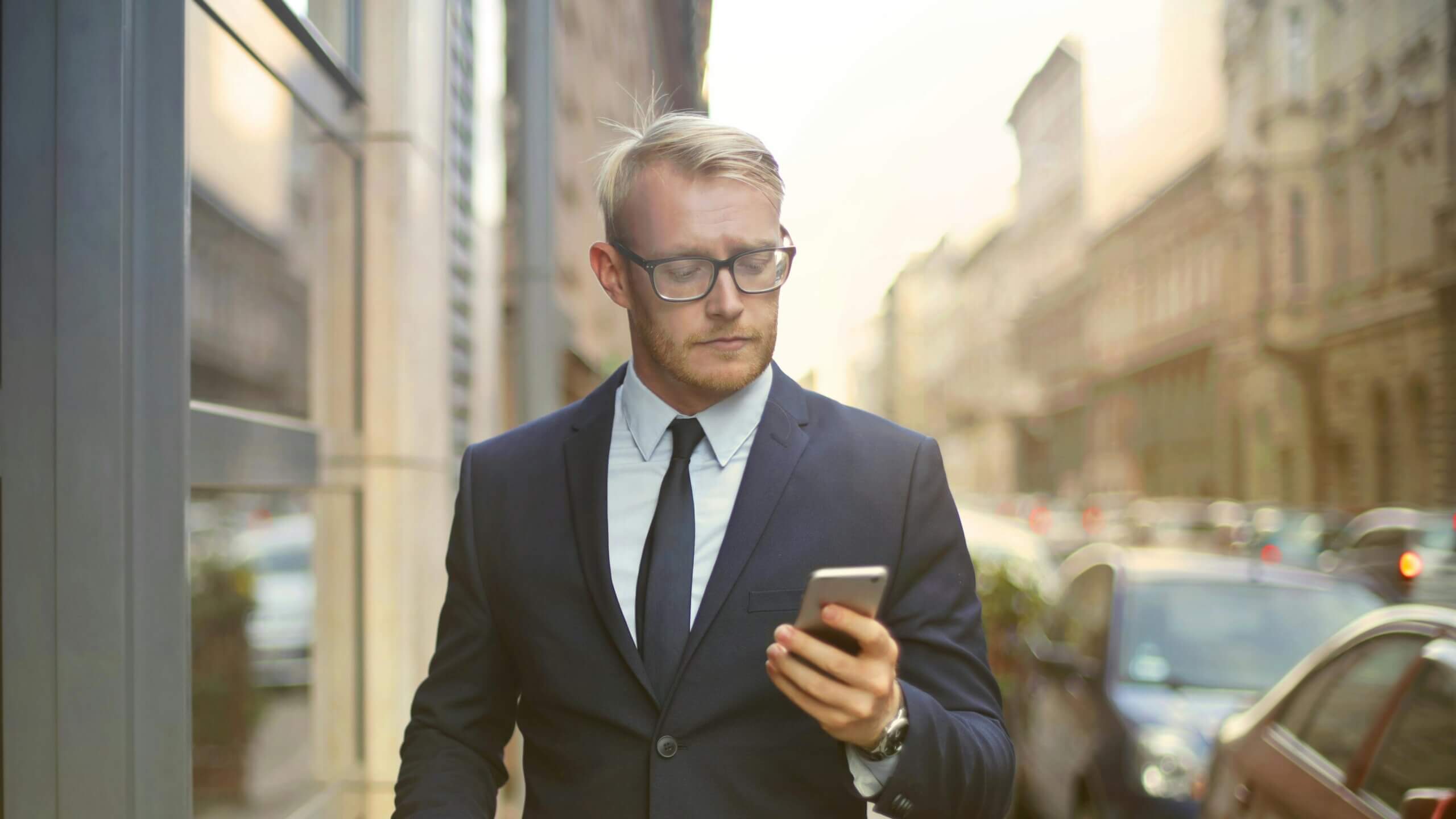 Blond man in glasses and suit looking at smartphone on a city street with cars in the background